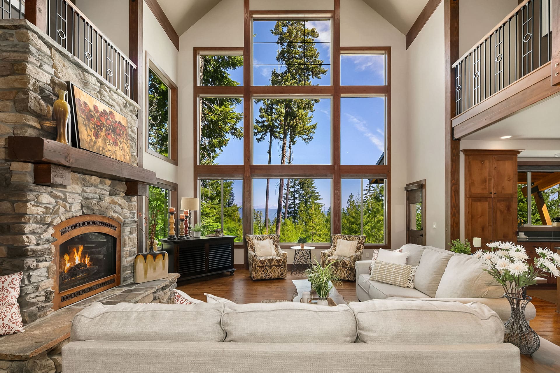 A spacious living room with a high ceiling and a large window offering a forest view. It features a stone fireplace, two armchairs, a sofa, a coffee table with flowers, and a console with lamps. The interior has a rustic yet modern style with wooden accents.