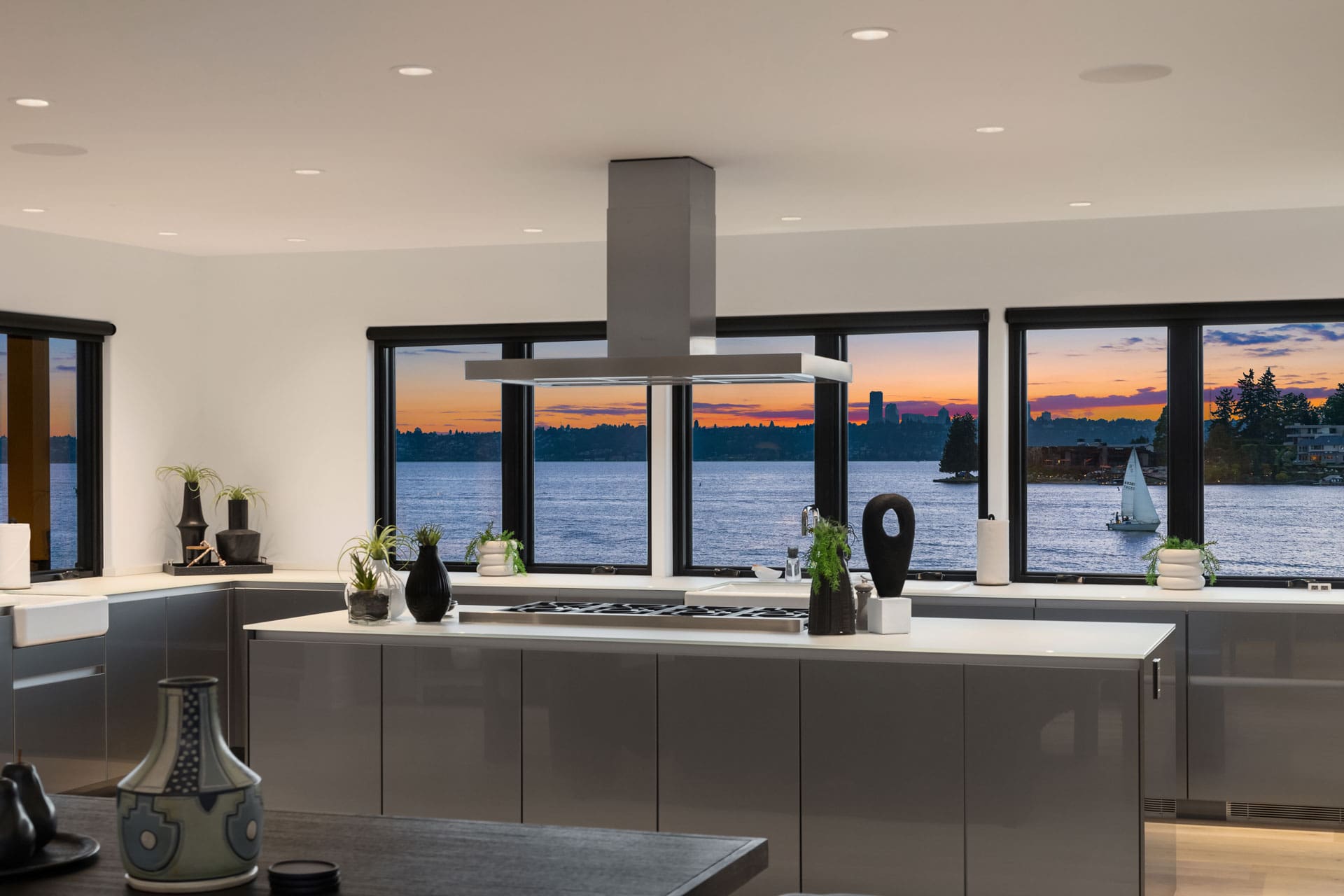 A modern kitchen with sleek gray cabinets, a large island, and a minimalist design. Large windows reveal a picturesque sunset over a serene lake with sailboats, adding natural beauty to the contemporary interior. Various small plants and decorative items adorn the space.