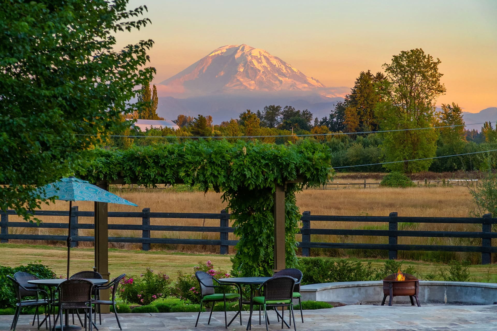 A serene outdoor patio with tables and chairs is set near a lush garden with an arbor covered in greenery. In the distance, a snow-capped mountain stands majestically against a pastel sky at sunset, providing a picturesque backdrop to the tranquil scene.