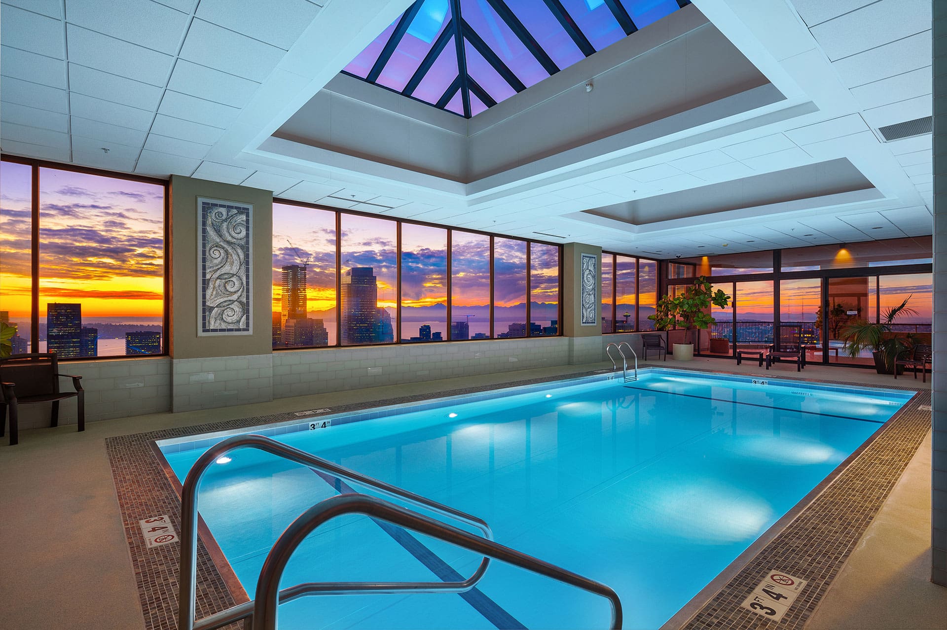 An indoor pool with clear blue water is surrounded by modern decor and large windows revealing a vibrant cityscape at sunset. The ceiling features a skylight, and the pool area is adorned with potted plants and chairs. The water depth marks are visible on the pool edge.