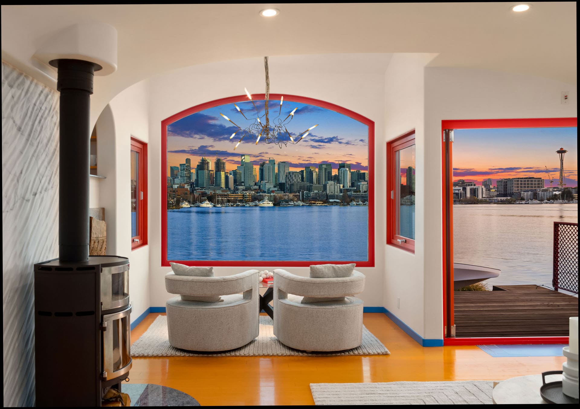 A modern room with two white armchairs facing a large window overlooking a city skyline at sunset. The window has a vivid red frame, and nearby is a wood-burning stove. The room features white walls, a decorative chandelier, and a view of a body of water.