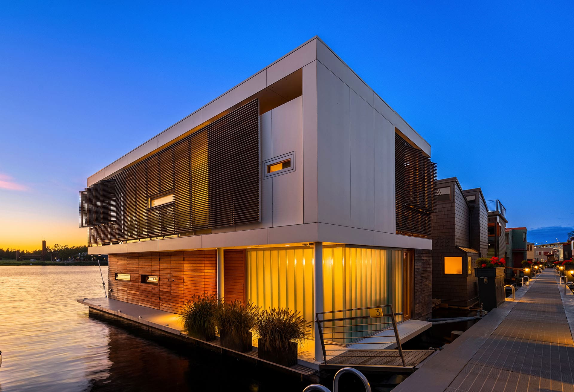 A modern, two-story houseboat sits on the water at sunset. The sleek design features large windows, wooden slats on the second floor, and soft lighting illuminating the ground floor. The houseboat is docked alongside a walkway with potted plants and a clear evening sky above.