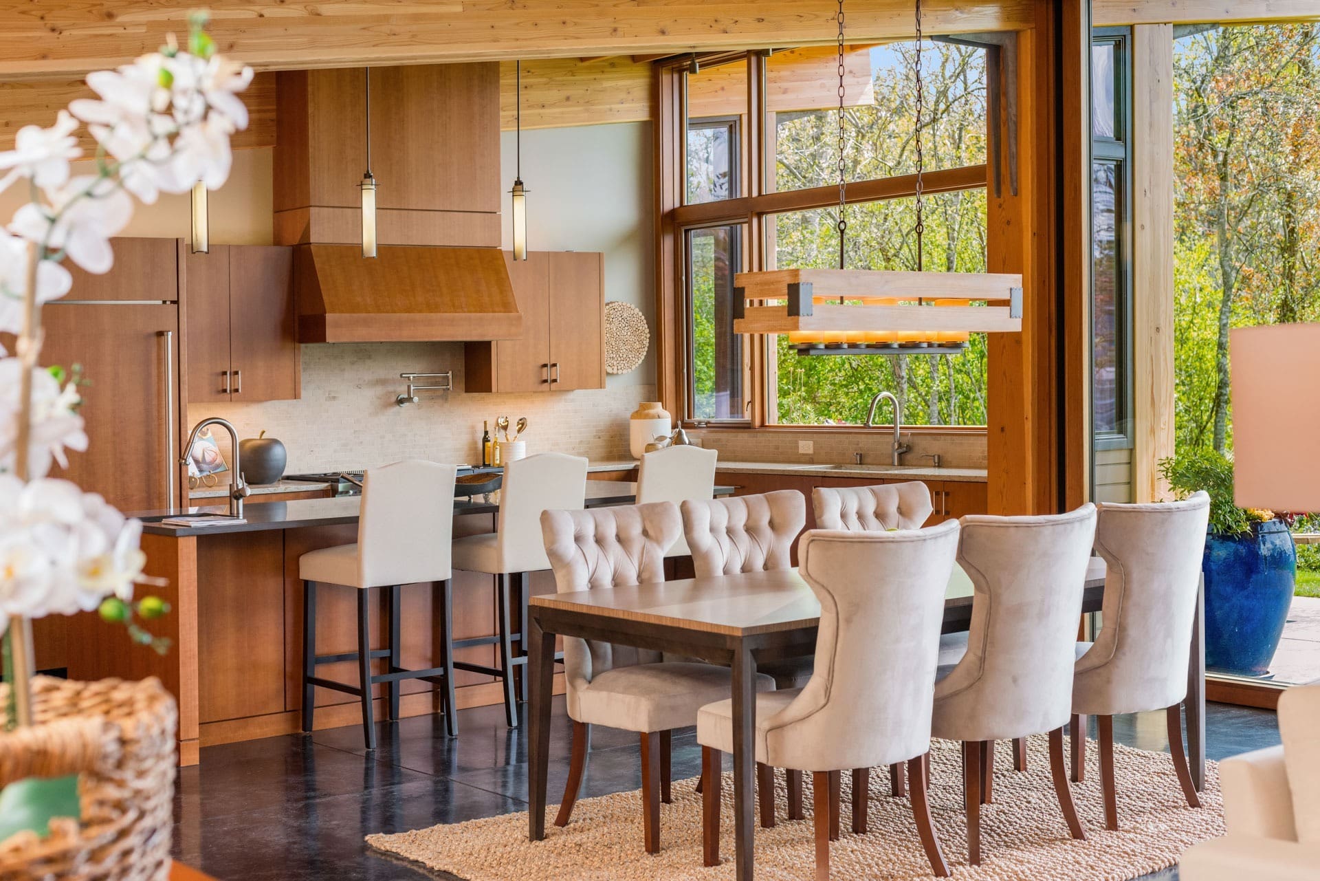 A modern kitchen and dining area with wooden cabinets and large windows. The kitchen includes a center island with white high chairs, while the dining table is surrounded by upholstered chairs. A woven rug lies beneath the dining set, and a large plant is visible.