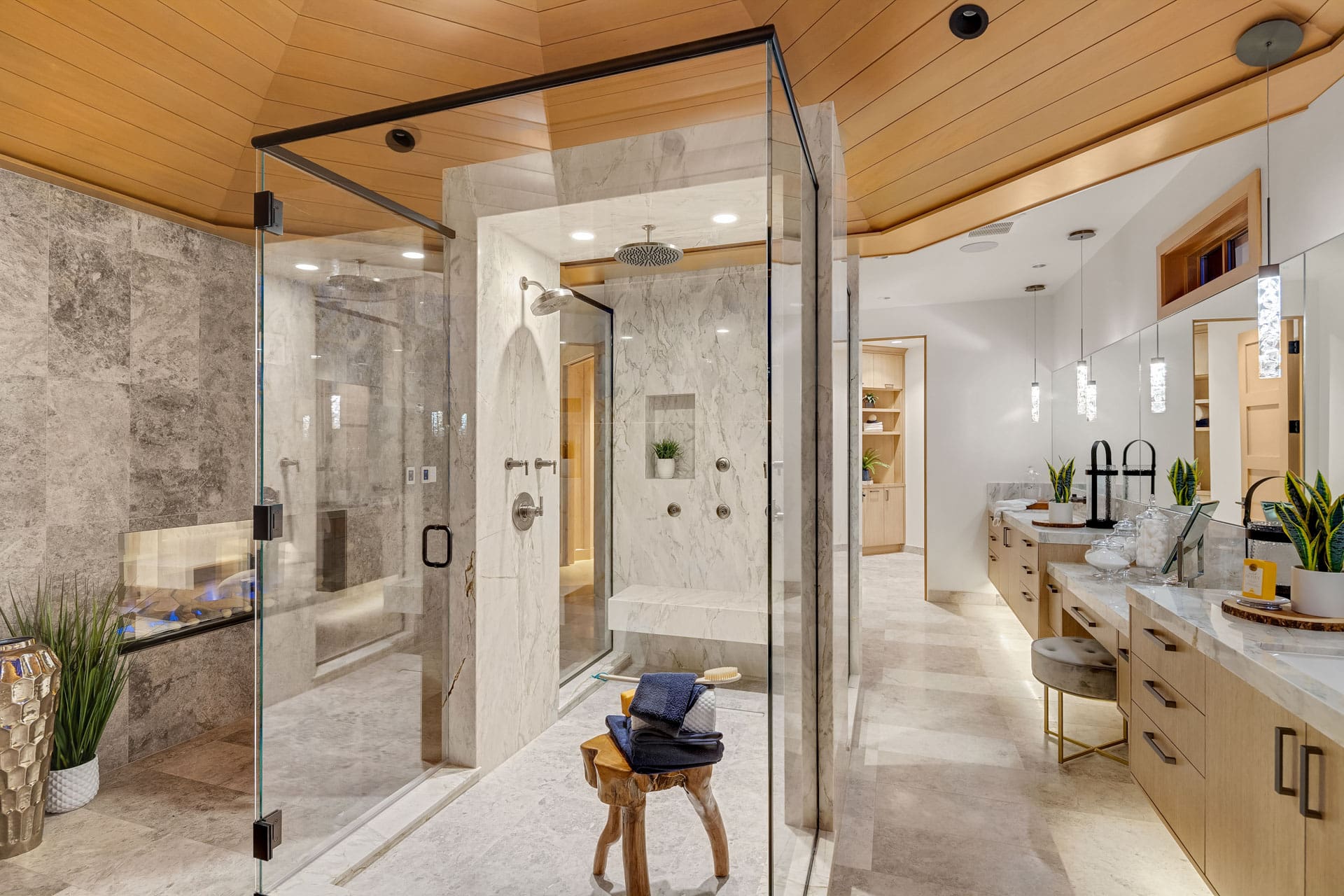 A luxurious bathroom with a glass-enclosed shower, stone tiles, and a wooden ceiling. The shower features multiple showerheads and a built-in bench. There is a dual-sink vanity with plants and decor, and a towel is neatly placed on a wooden stool in the center.