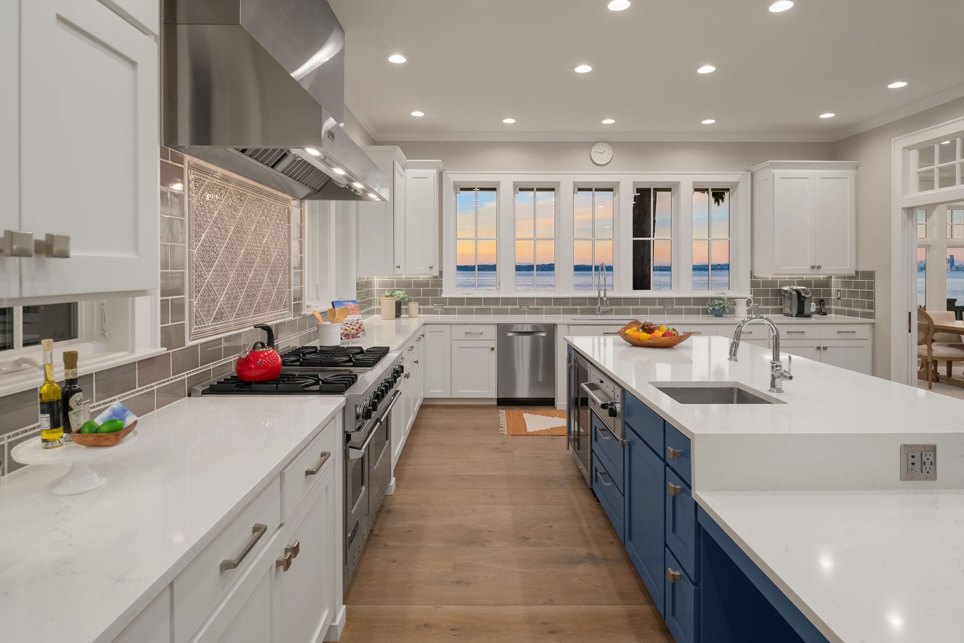 Modern kitchen with white cabinets, gray tile backsplash, and stainless-steel appliances including a large range hood and double oven. Island with blue cabinets and white countertops. Windows showcase a sunset view over the water. Wood flooring throughout.