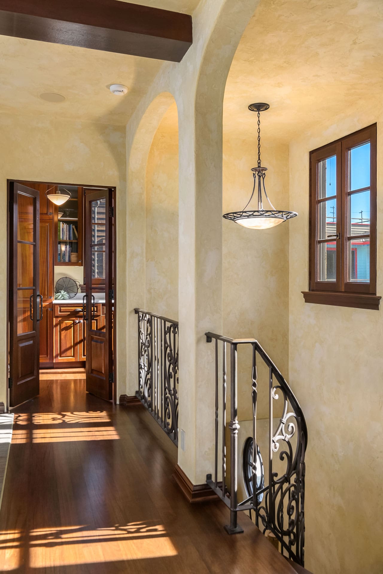 A bright hallway with beige walls and wooden flooring leads to a room in the distance with shelves full of books. Ornate iron railings line a staircase on the right. An elegant hanging light fixture illuminates the space, and exposed beams enhance the ceiling.