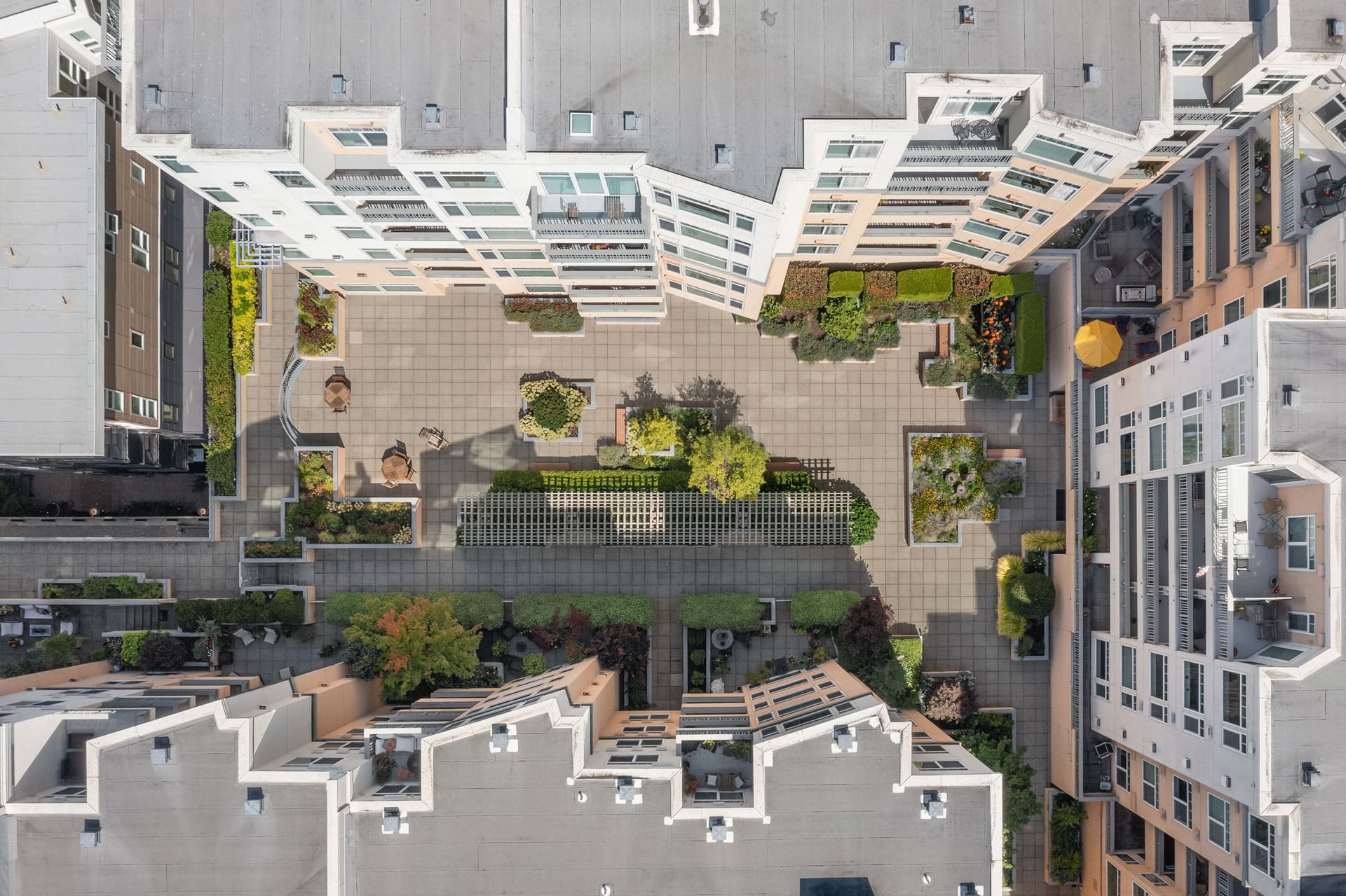 Aerial view of a rectangular courtyard surrounded by modern apartment buildings. The courtyard features paved walkways, green spaces, trees, and shaded seating areas. The buildings have balconies overlooking the courtyard.