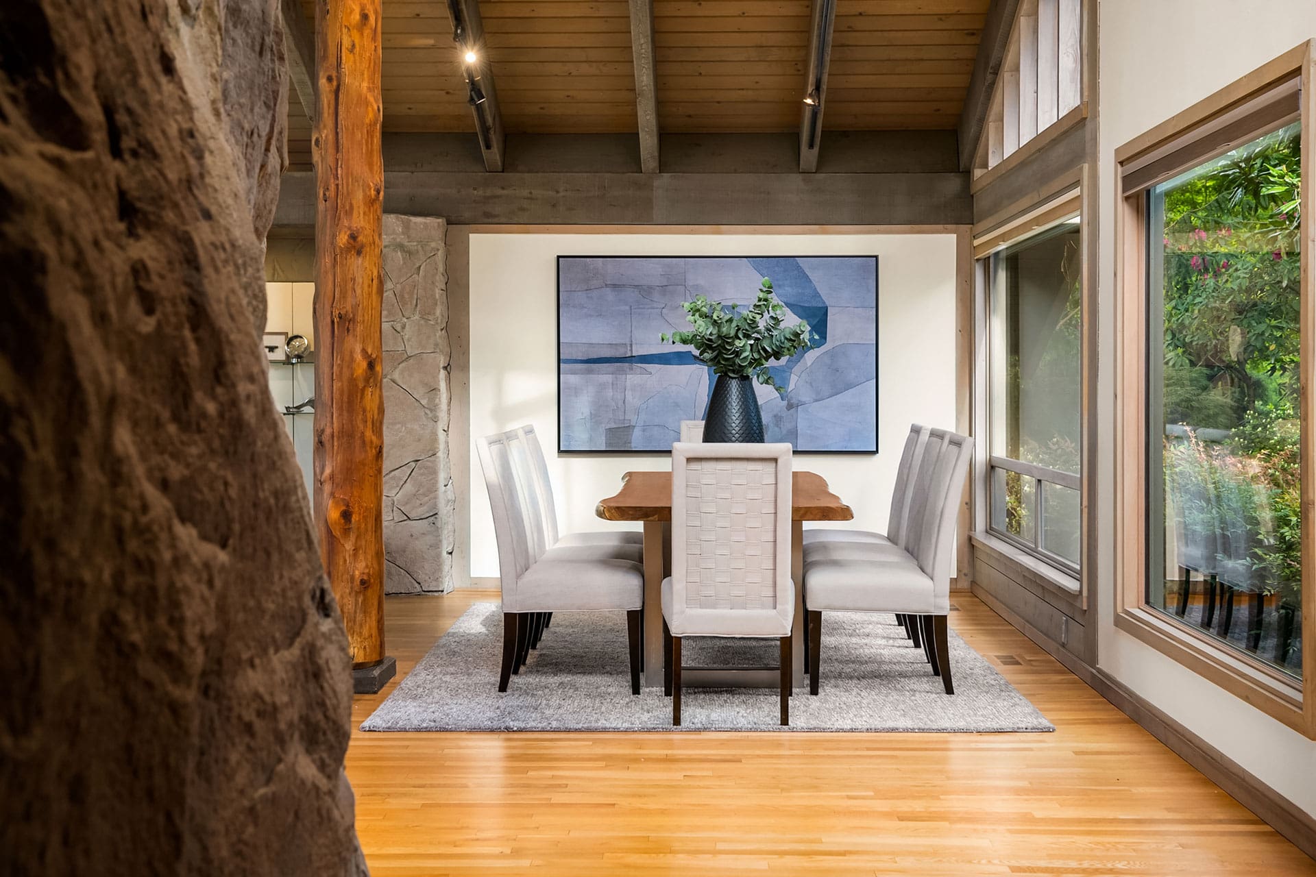 A modern dining room with a wooden table and six white upholstered chairs on a gray rug. A large abstract painting hangs on the wall. Tall windows let in natural light, and an exposed wooden beam adds rustic charm. A vase with greenery is in the center of the table.