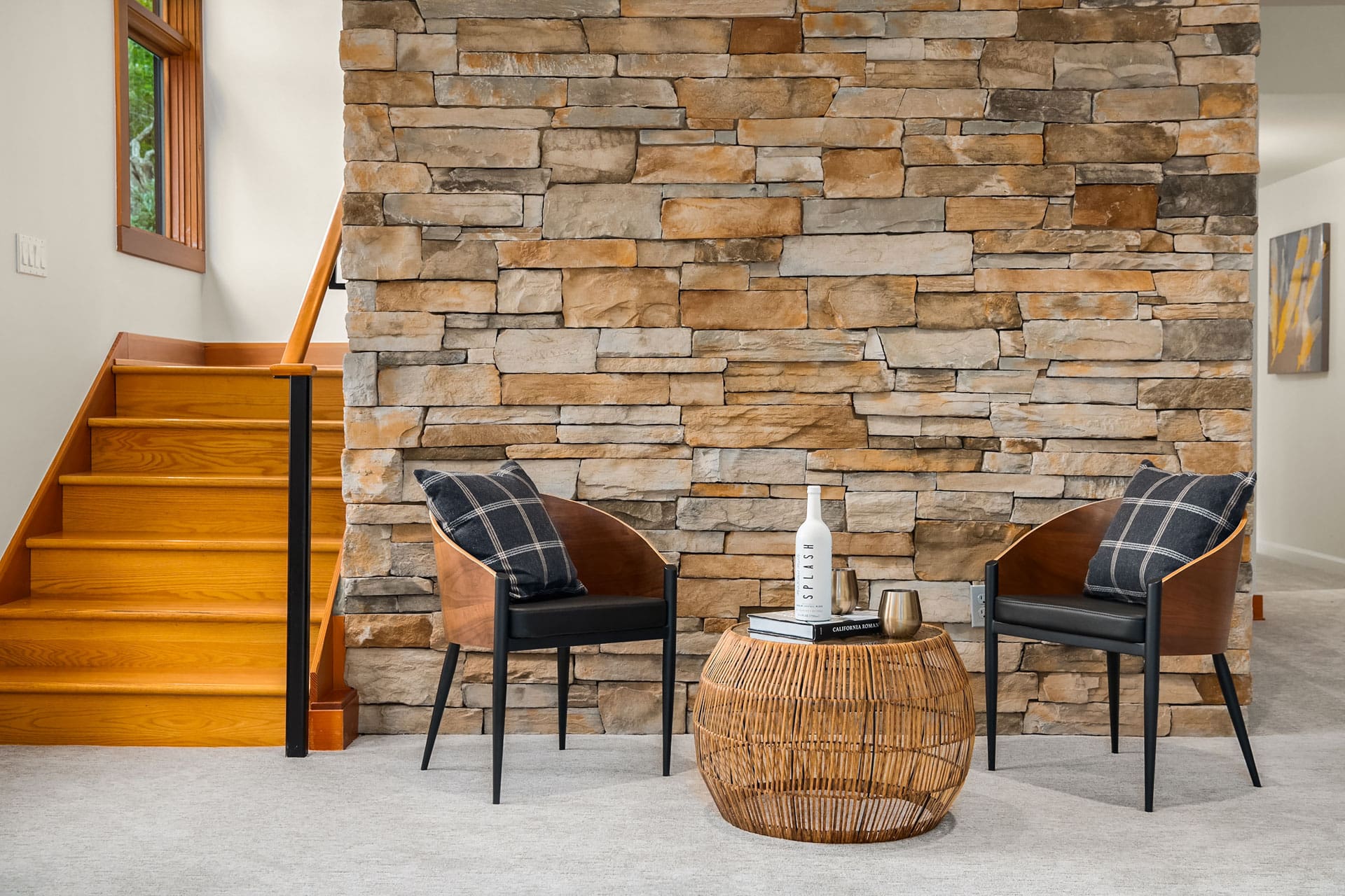 Modern interior with a rustic stone wall, wooden staircase on the left, two cushioned wooden chairs, and a round wicker coffee table in the center. Table holds a white vase, books, and a candle. Room has minimalistic decor and warm lighting.