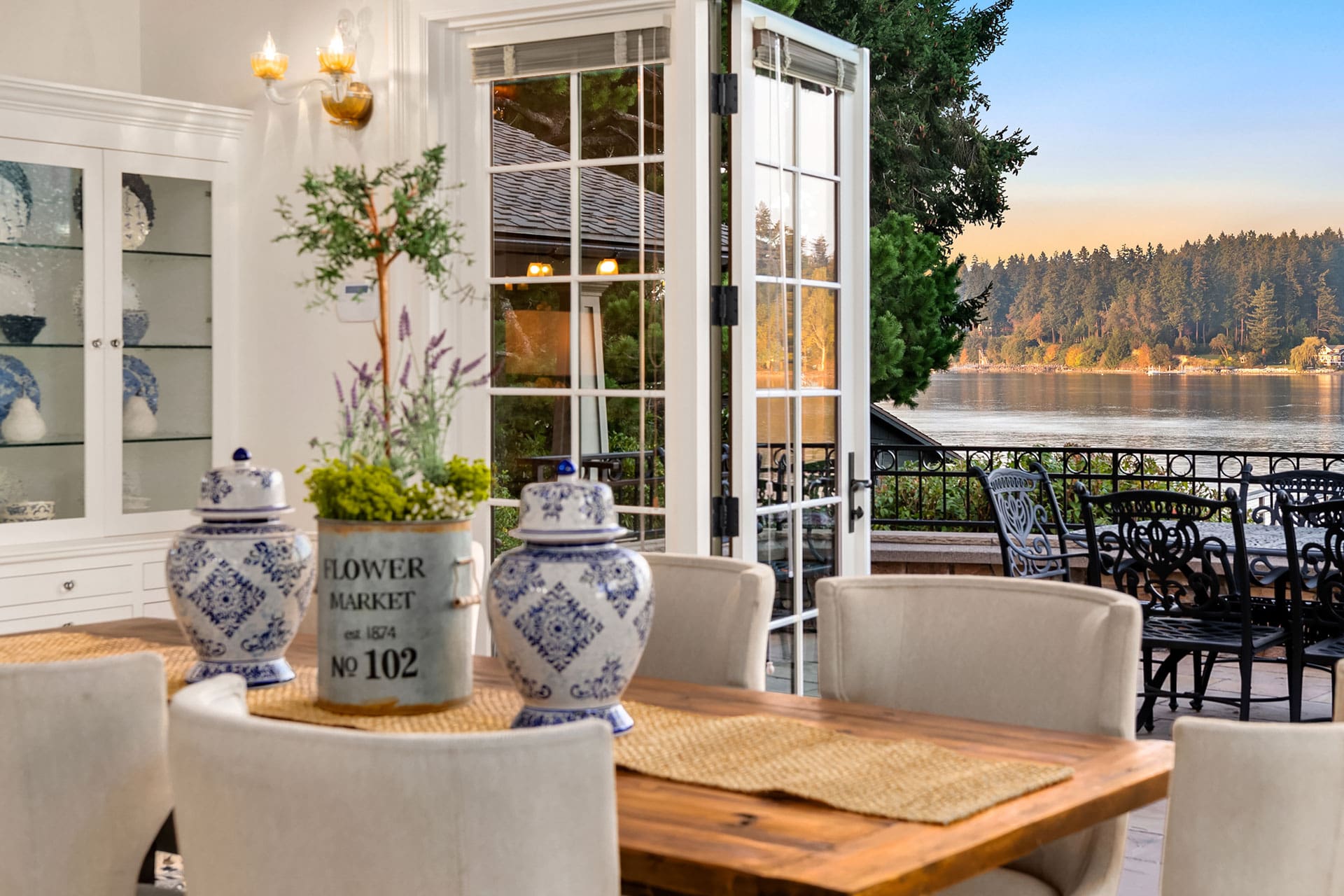 A dining room with a wooden table, cream-colored chairs, and blue and white porcelain decor. Double doors open to a patio, showcasing a serene lake and forested landscape at sunset. The room has a cozy, elegant design with outdoor views enhancing its charm.