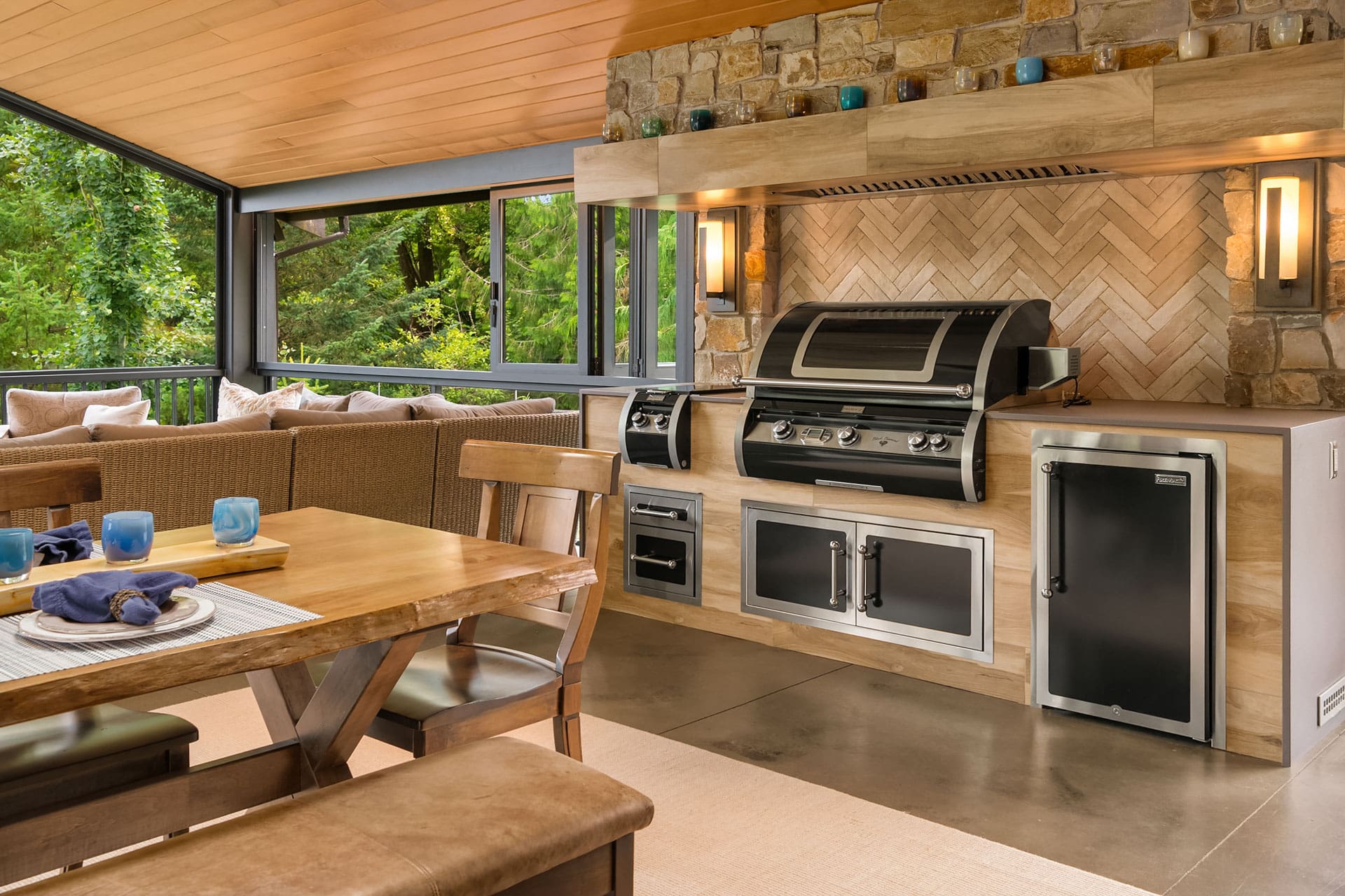 A modern outdoor kitchen with a stainless steel grill and appliances set in a wooden cabinet against a stone wall. The space includes a wooden dining table, cushioned bench seating, and large windows showcasing lush greenery, creating an elegant and inviting atmosphere.