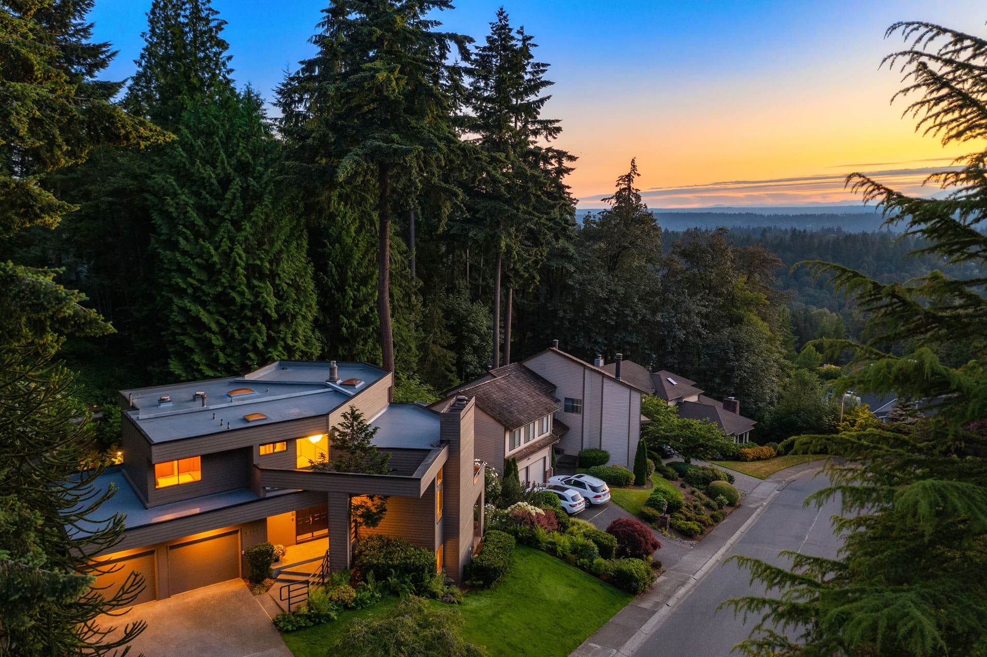 An aerial view of a residential neighborhood featuring modern houses nestled among tall trees. One house has illuminated windows and a well-maintained lawn. The scene is set during sunset, with a colorful sky in the background and a serene, wooded landscape.