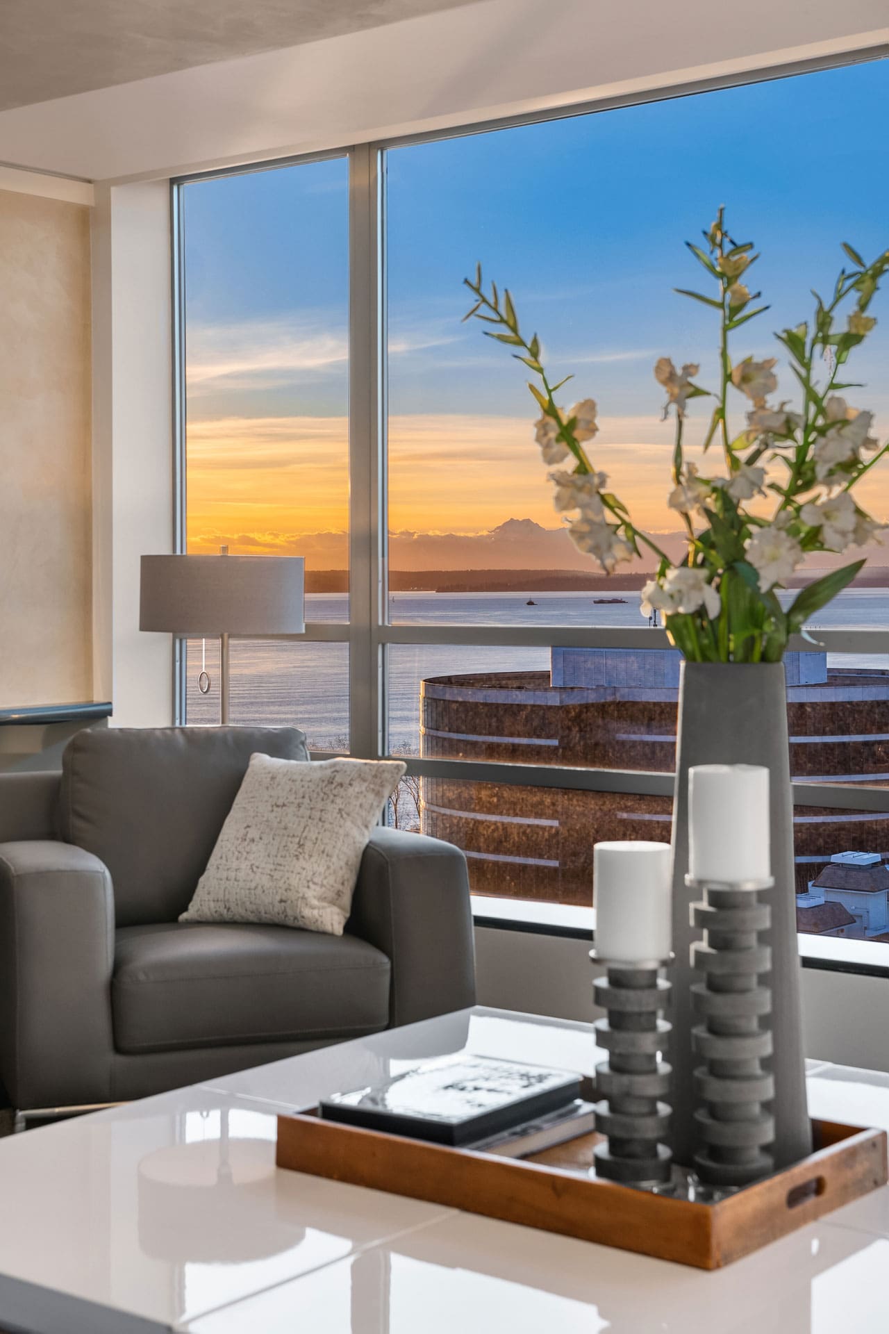 Modern living room with a gray armchair, a lamp, and a side table holding a vase with white flowers. A white coffee table features a tray with two candles and a book. Large windows offer a view of the ocean and sunset.