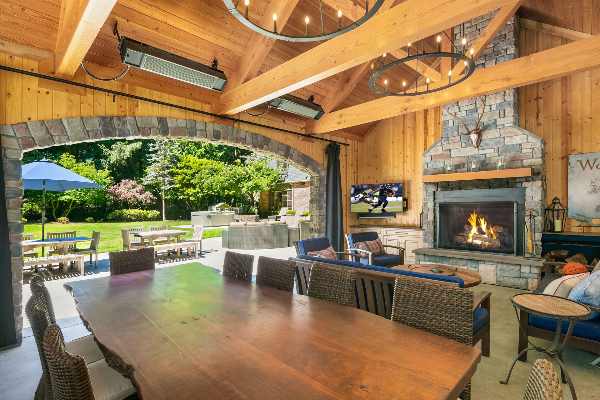 A spacious outdoor patio with a wooden dining table and wicker chairs. The area features a stone fireplace with a lit fire, a TV mounted above it, and a vaulted wooden ceiling with large chandeliers. Beyond the patio, there's a lush green garden with additional seating and an umbrella.