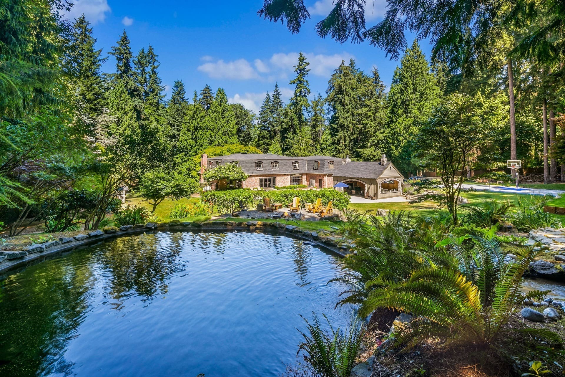 A large brick house with a shingled roof is nestled among tall trees. In the foreground, there is a serene pond surrounded by lush greenery. Near the pond, there are patio chairs and a stone pathway leading to the house. The sky is bright and clear.