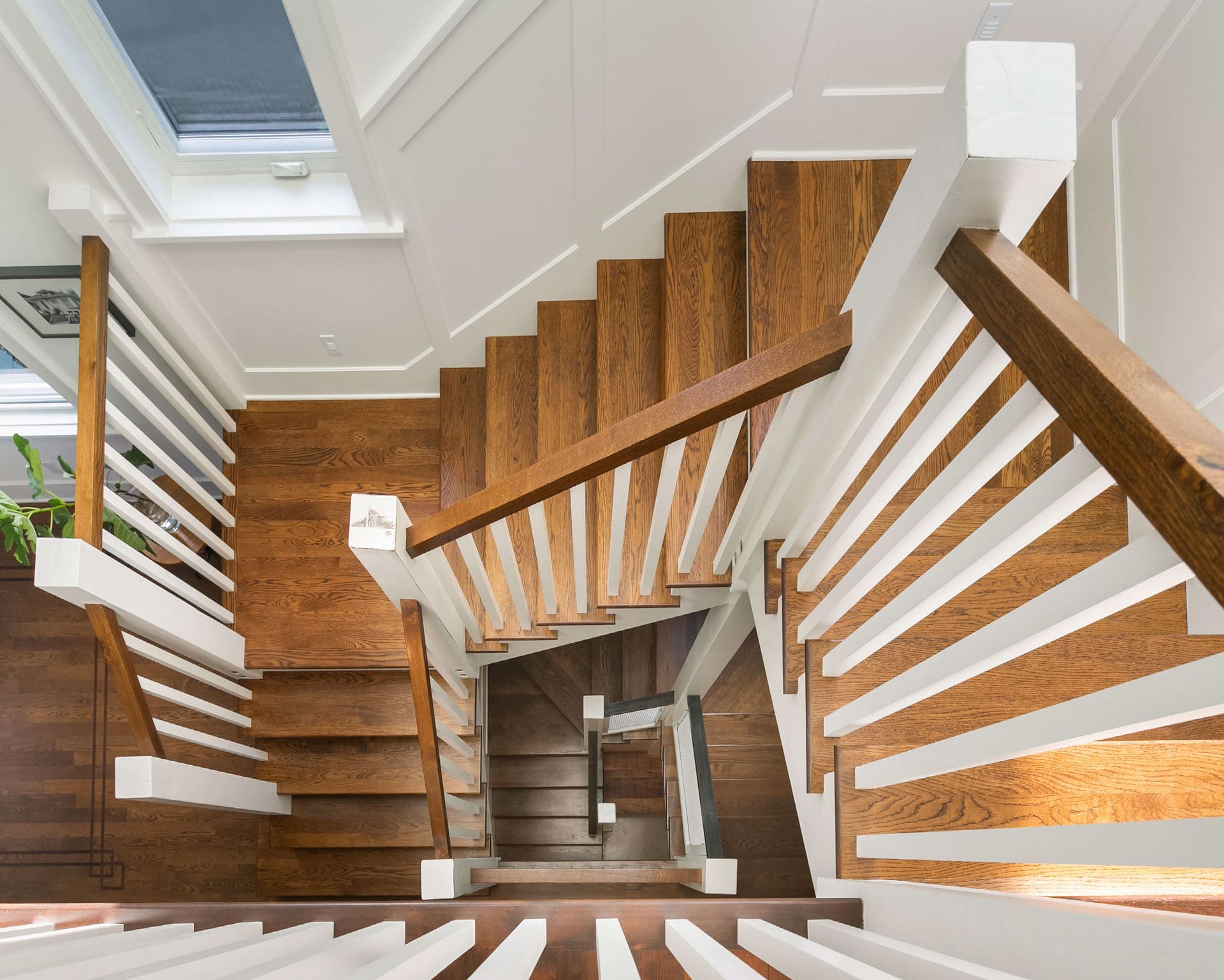 An aerial view of a wooden staircase with white railings in a modern, well-lit interior. The stairs lead down in a geometric pattern, creating a visually appealing spiral effect. The walls are white, and a window can be seen in the upper left corner.