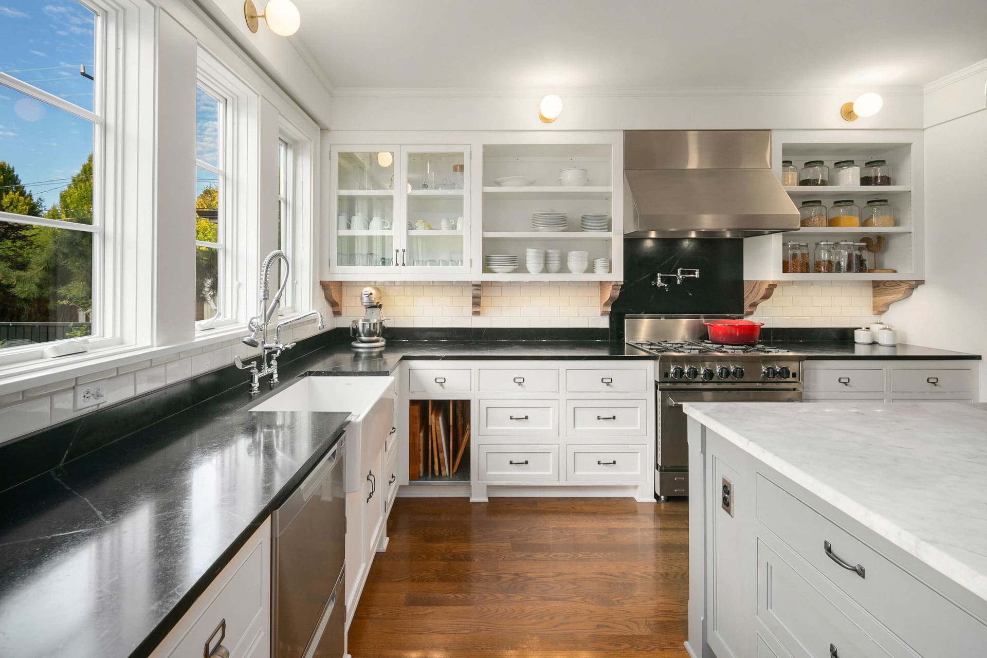 A bright, modern kitchen with large windows. It features black countertops with white cabinets and drawers. A stove with a stainless steel hood is visible, with shelves and utensils on the wall. A kitchen island with a white countertop sits in the center.