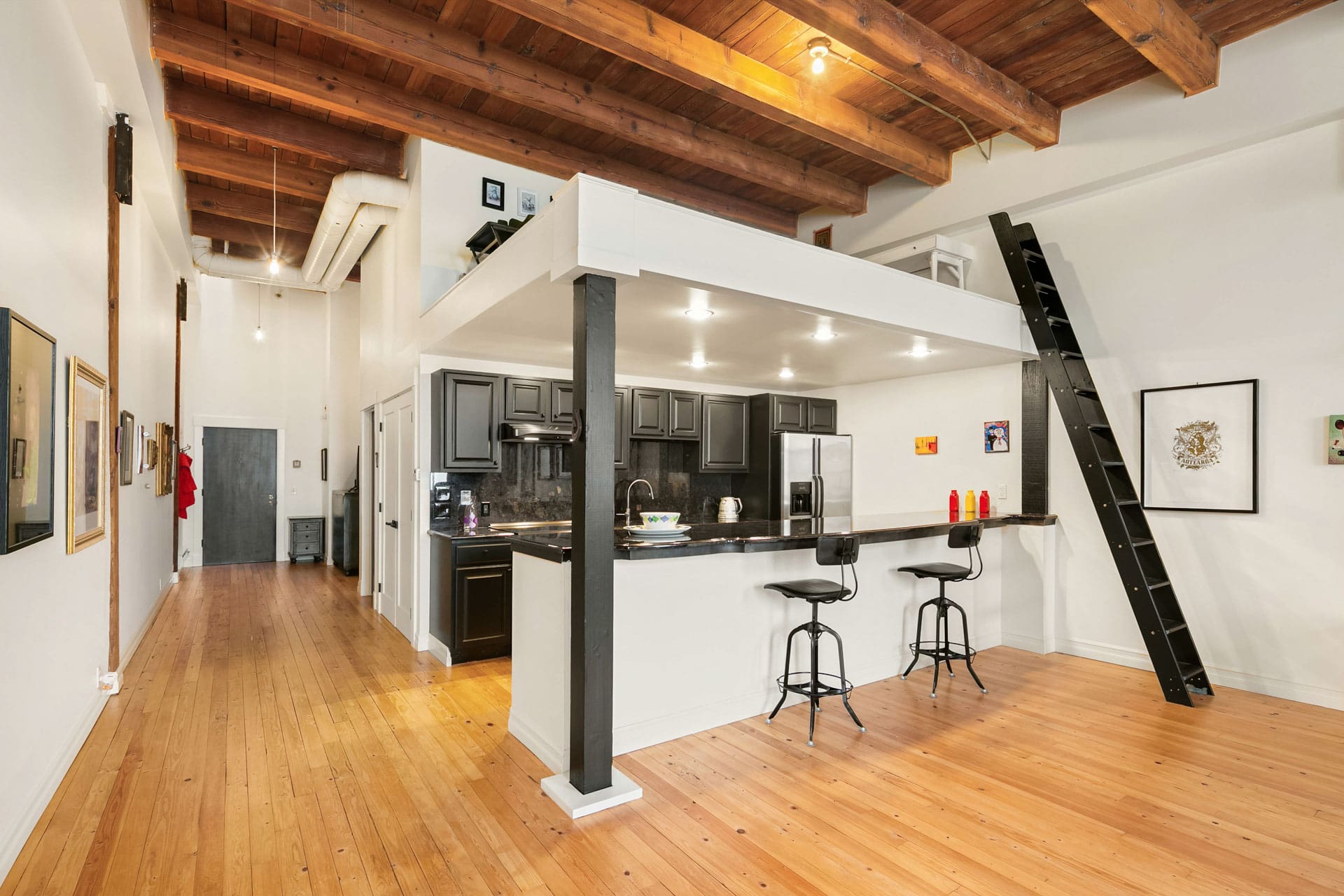 A modern loft-style kitchen with dark cabinets, a high ceiling with exposed wooden beams, and a small breakfast bar with two stools. The open space includes wood flooring and a ladder leaning against the wall leading up to a lofted area.