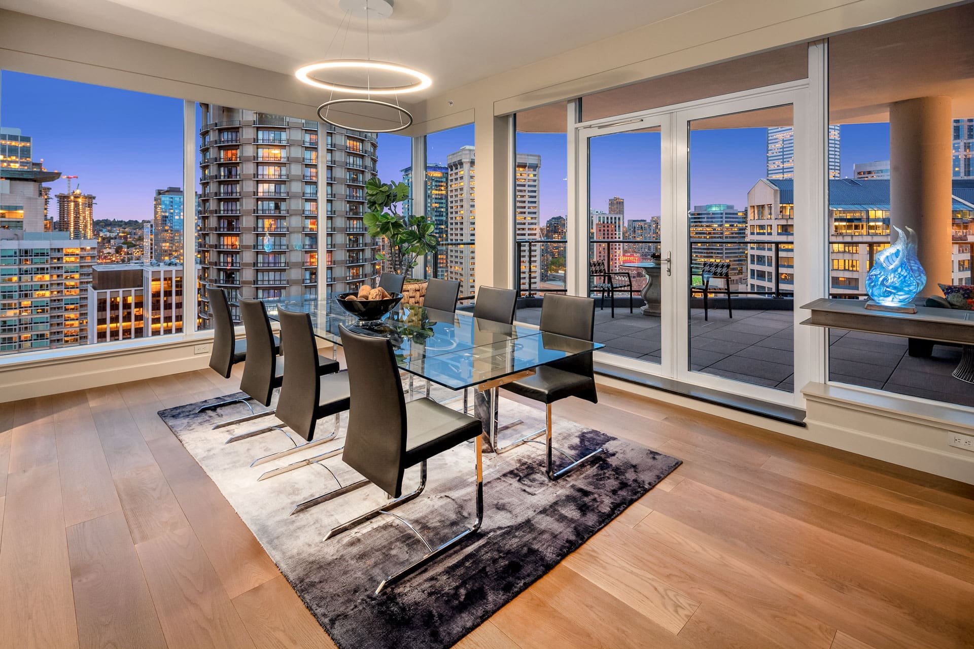 A modern dining room with a glass table and eight black chairs on a stylish rug. Floor-to-ceiling windows offer a stunning view of a cityscape at dusk. A circular chandelier hangs above the table, and a balcony is accessible through sliding glass doors.