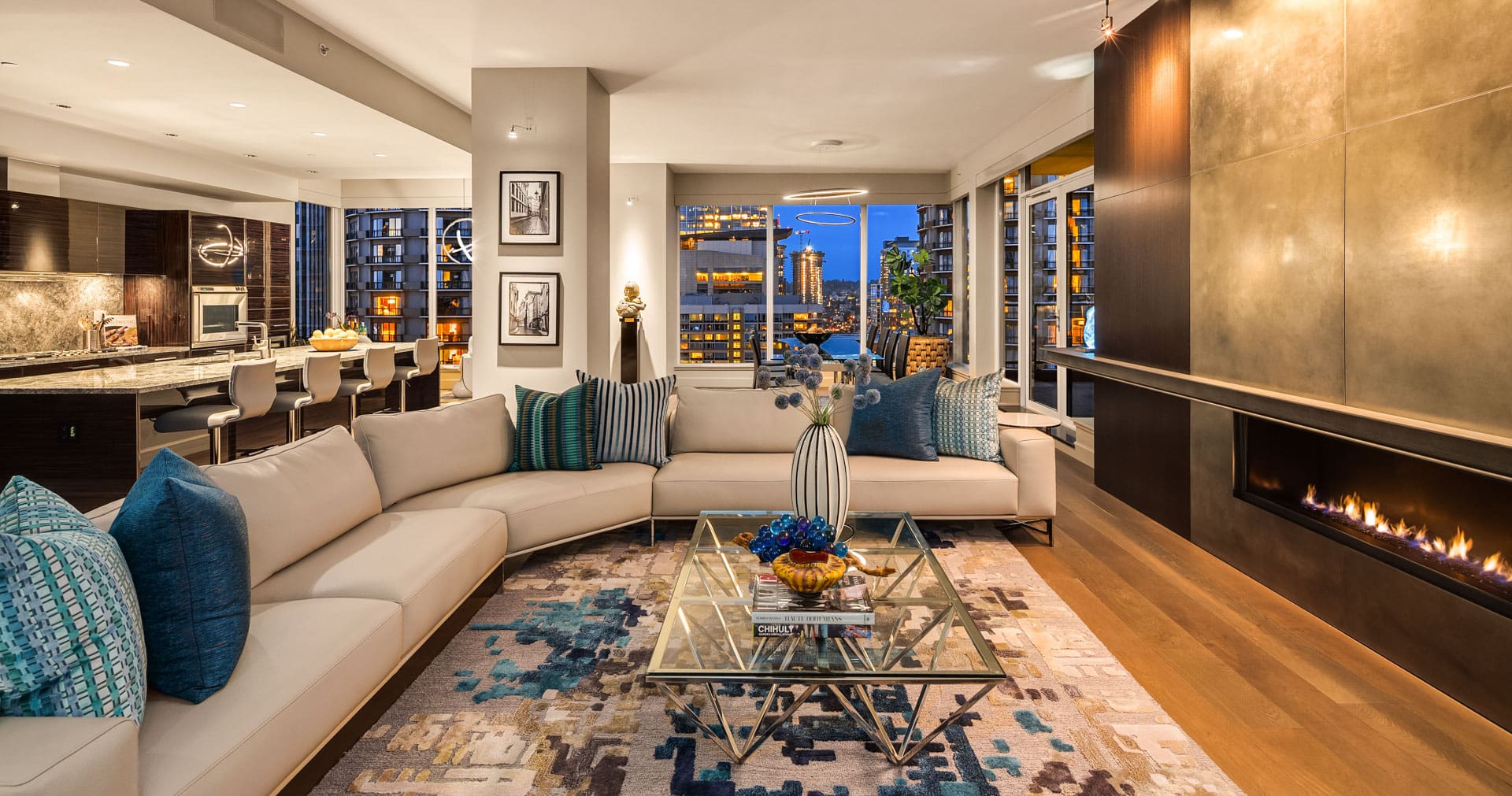 Luxurious modern living room with a large cream sectional sofa adorned with blue and green accent pillows around a glass coffee table on a colorful rug. The room features a contemporary fireplace, open kitchen, and cityscape view through large windows.