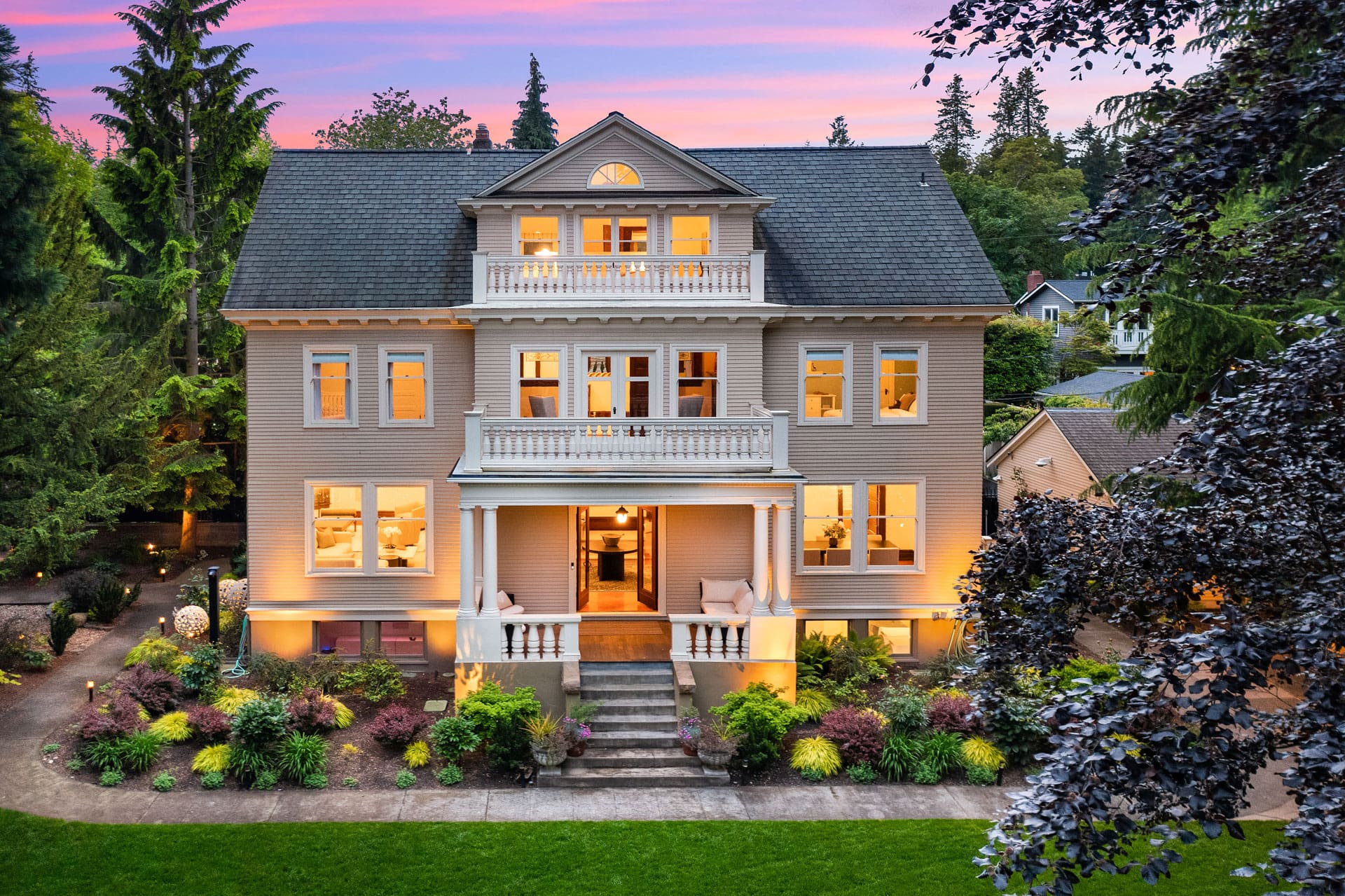 A large, three-story house with a classic architectural style, illuminated warmly at dusk. It features a symmetrical facade with a central balcony, multiple windows, and a welcoming front porch. The surrounding garden is well-maintained, with trees and shrubs.