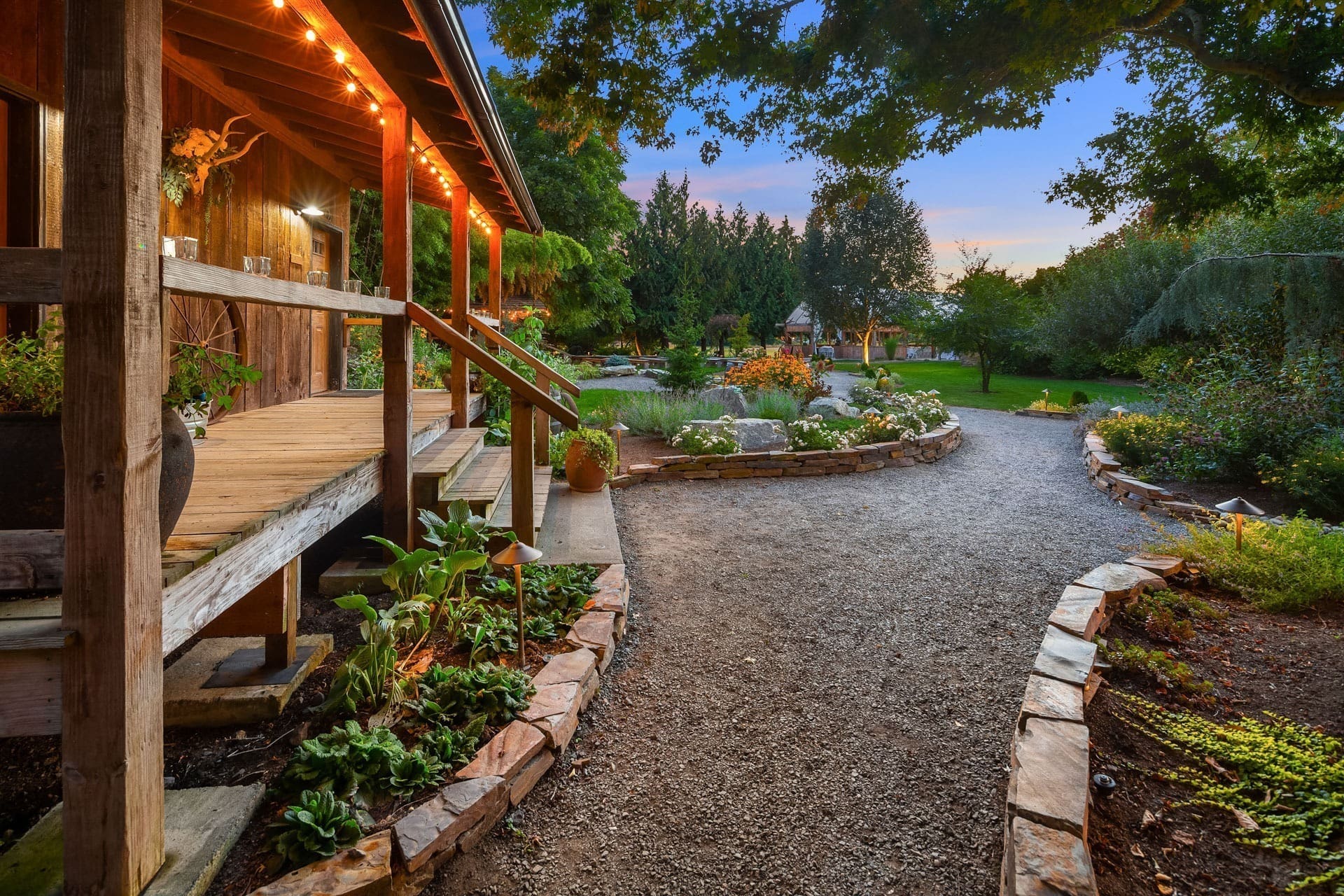 A cozy wooden porch with string lights overlooks a scenic garden at sunset. The garden path, lined with plants and stone borders, curves gently, leading to a green lawn and trees in the background. Various potted plants and flowers adorn the garden.