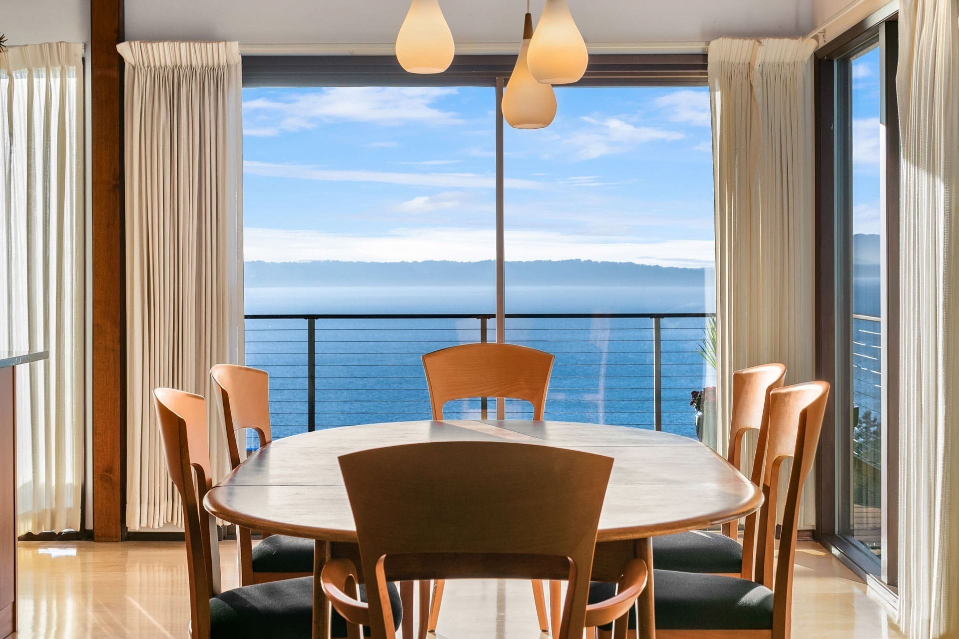 A modern dining room with large windows providing a scenic ocean view. It features a round wooden table surrounded by six wooden chairs with black cushions. Pendant lights hang above the table, and white curtains frame the windows, letting in ample natural light.