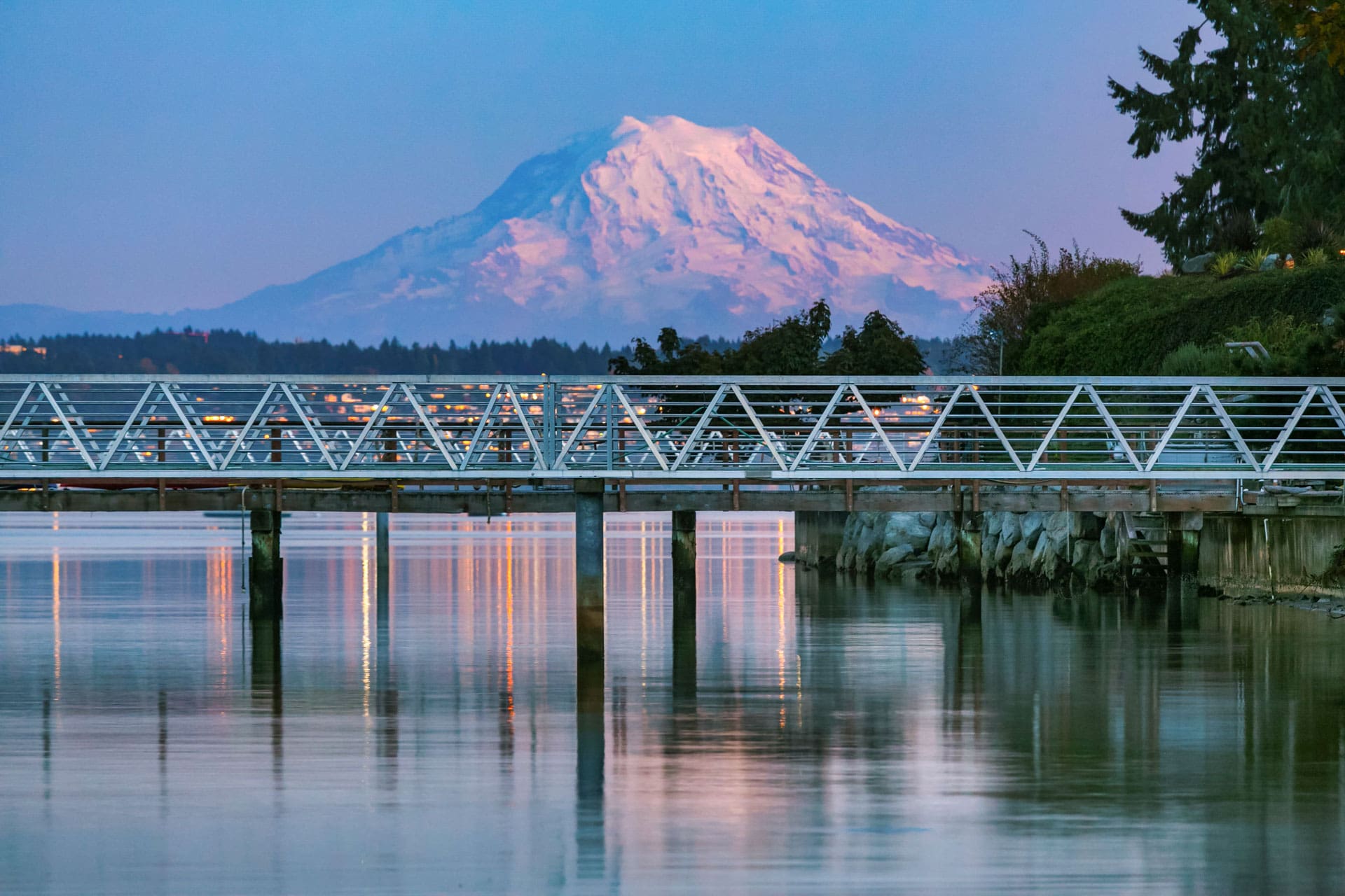 A scenic view of a snow-capped mountain at sunset, seen across a calm body of water. A long, metal pedestrian bridge extends over the water, with trees and small buildings in the background. The sky is a gradient of blue to purple, reflecting off the water's surface.