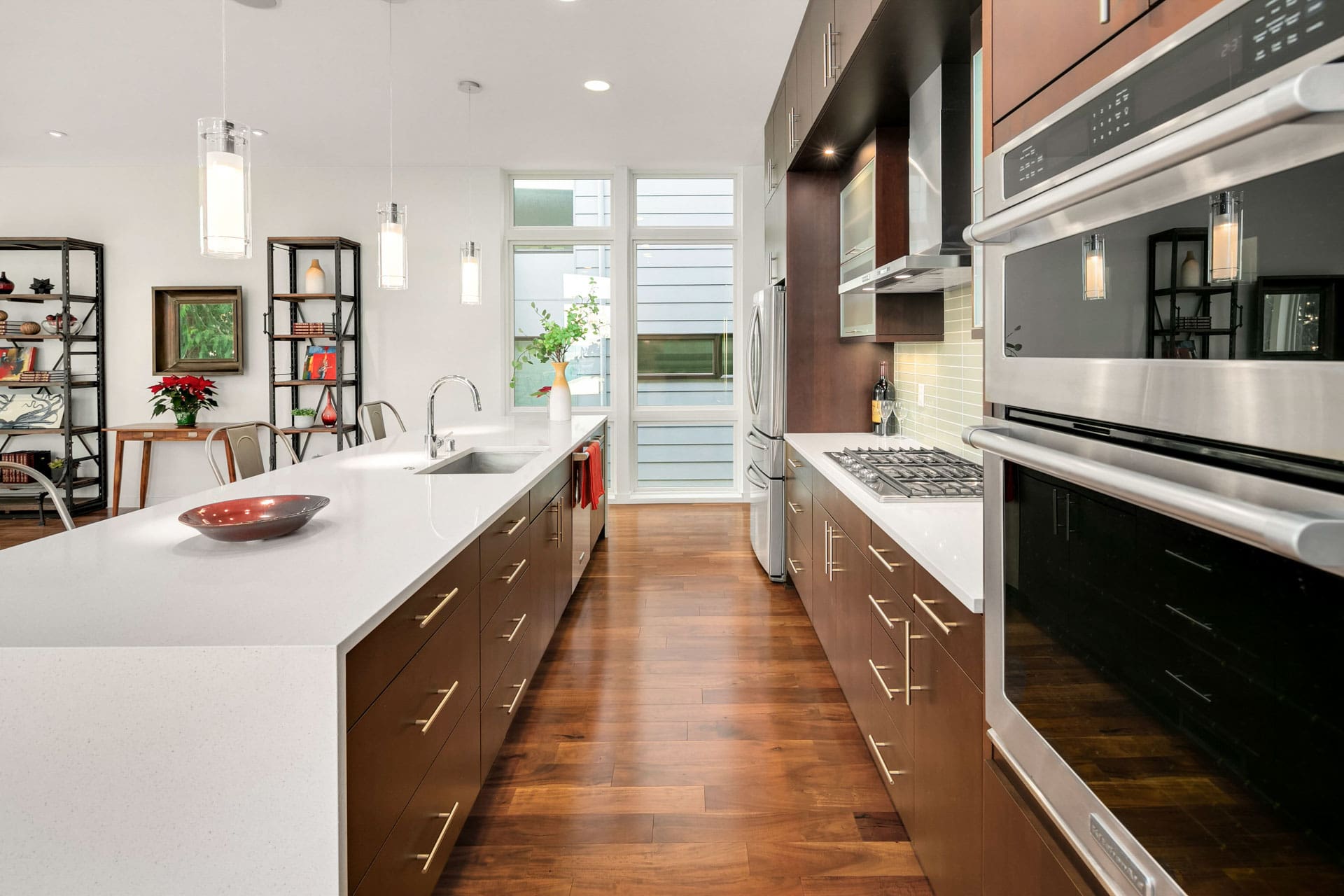 Modern kitchen with dark brown cabinets and stainless steel appliances, including a built-in oven and gas stovetop. A white countertop island with a sink and bar stools sits in the center. Bright pendant lights hang from the ceiling, and a large window offers ample natural light.