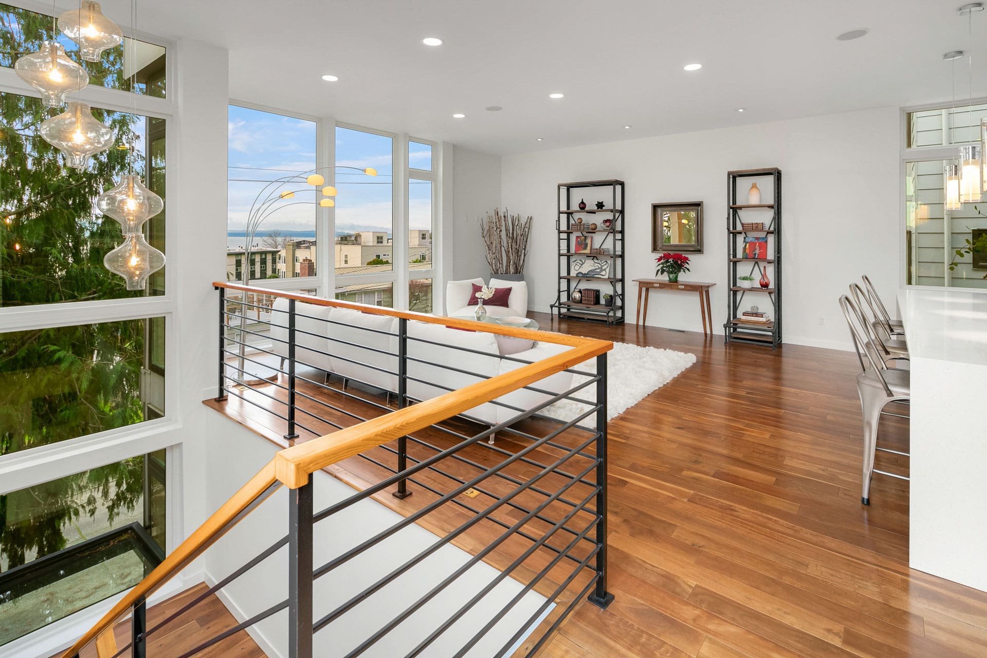 Modern, open-concept living area featuring large windows with a scenic view, wooden flooring, a white sofa, shelves with decor, and a dining area with metallic chairs. Stairs with a black and wooden railing lead from the lower level to the living space.