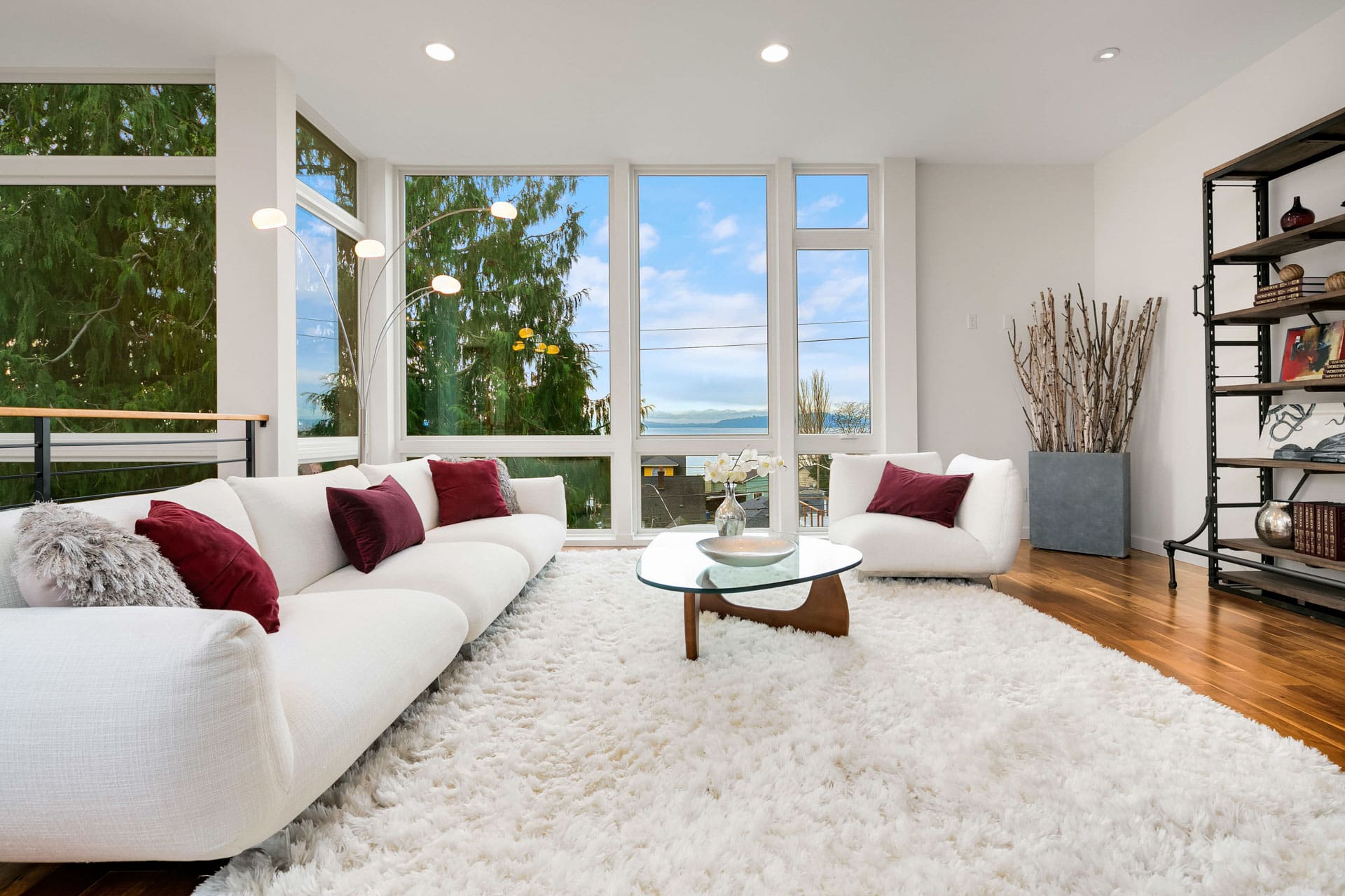 A modern living room with large windows showing an outdoor view. The room is furnished with a white sofa and a white lounge chair, both adorned with red and white pillows. There is a glass coffee table on a plush white rug and a tall shelf with decorative items.