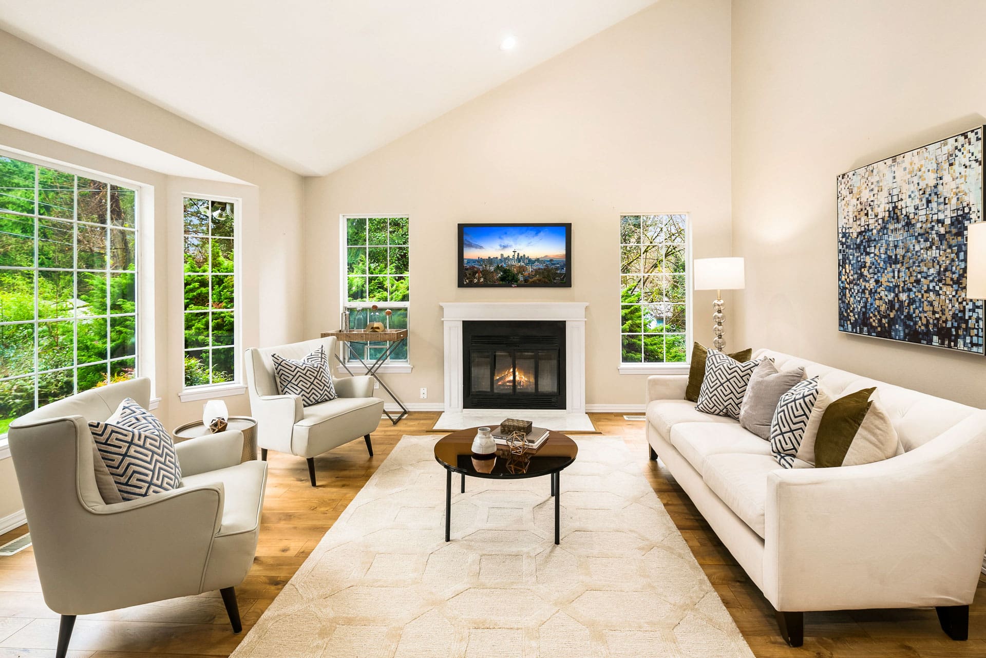 A bright, modern living room with a high ceiling, large windows, and light-colored walls. A white couch and two armchairs surround a black coffee table. A fireplace with a TV above it is centered on the back wall. The room features wooden floors and tasteful decor.