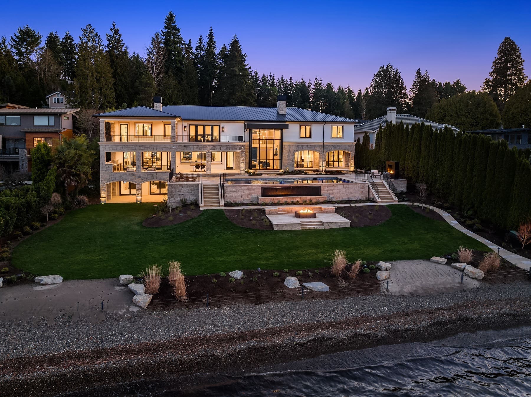 A luxurious two-story modern house with large windows and balconies, situated on a spacious waterfront lawn. The backyard features a patio with a fire pit, an elevated swimming pool, and landscaped gardens. Tall trees surround the property, which extends to a beach shoreline.