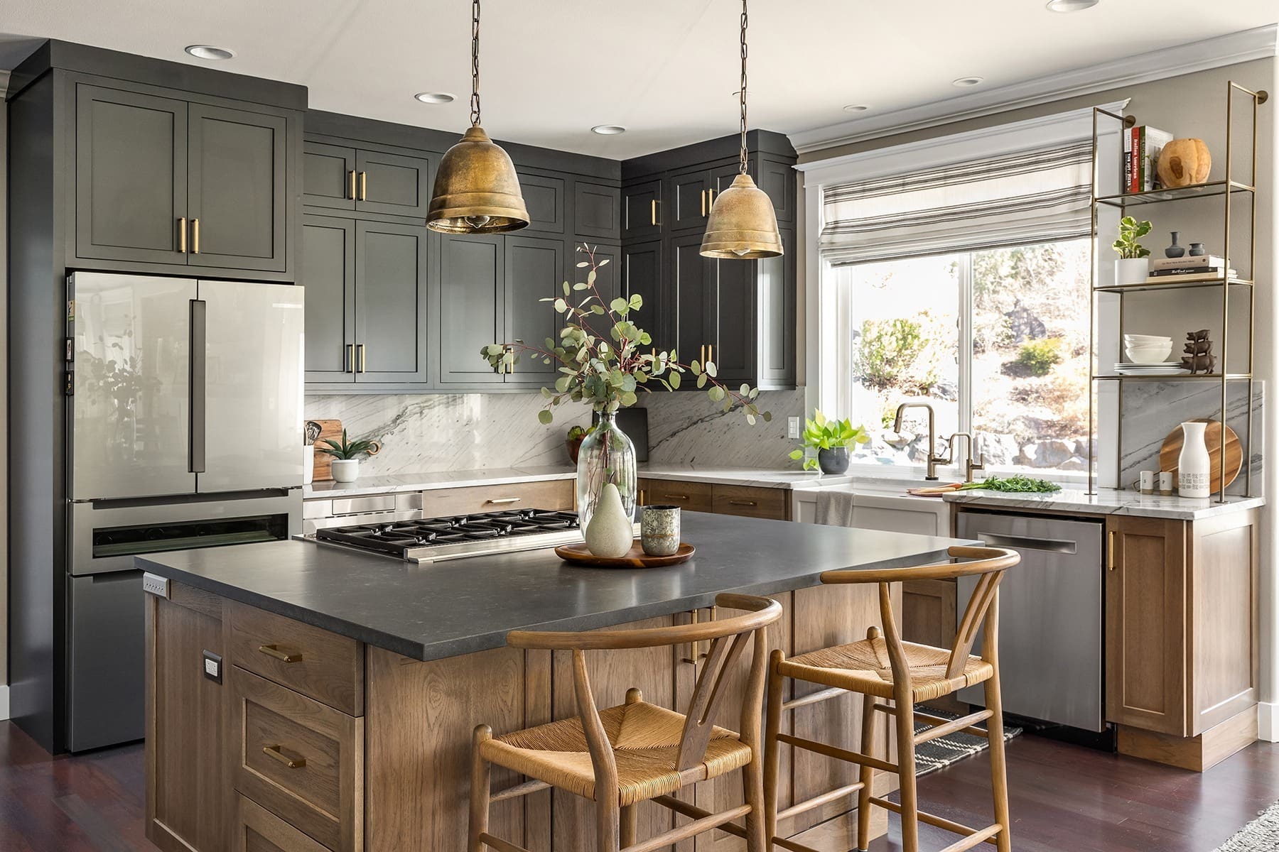 A modern kitchen features a large island with wooden base and black countertop, surrounded by wicker chairs. The refrigerator and cabinets are dark grey, while the backsplash and counters are white. Two bronze pendant lights hang above the island.