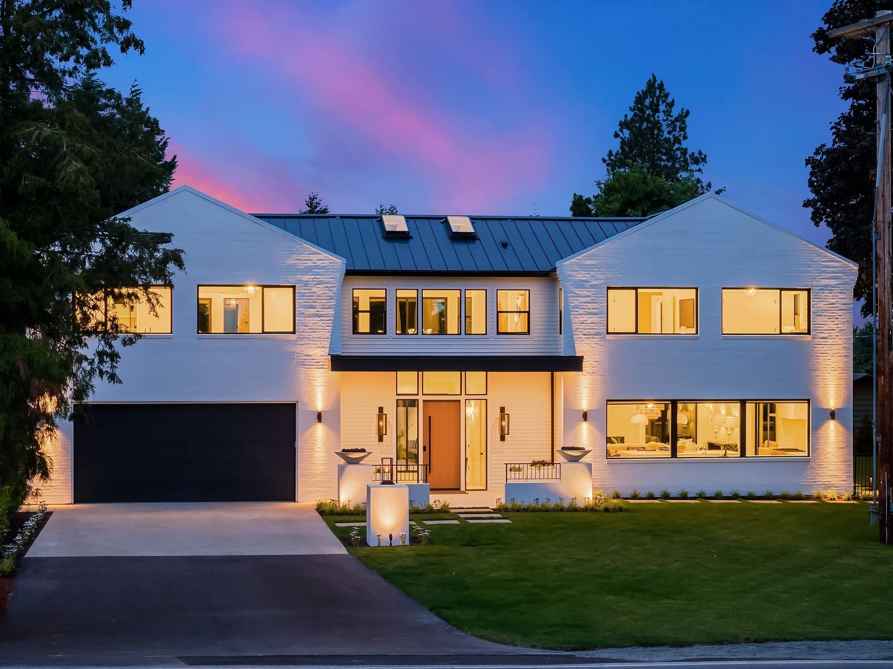 A modern two-story house with white brick exterior, large windows, and a black roof with skylights. The front is well-lit with warm lights, has a manicured lawn, and a wide driveway leading to a double garage. The sky is a vibrant mix of purple and pink hues at dusk.