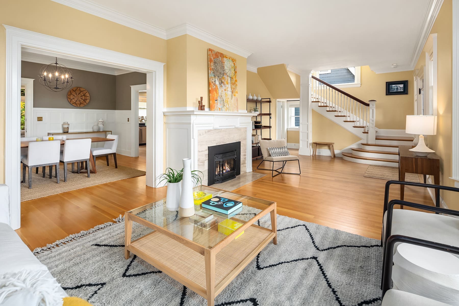 A spacious, elegant living room with light beige walls and wood flooring features a cozy fireplace with white trim. A wooden coffee table with books and plants sits on a patterned rug, and a staircase with white railing is visible in the background. A dining room is seen to the left.