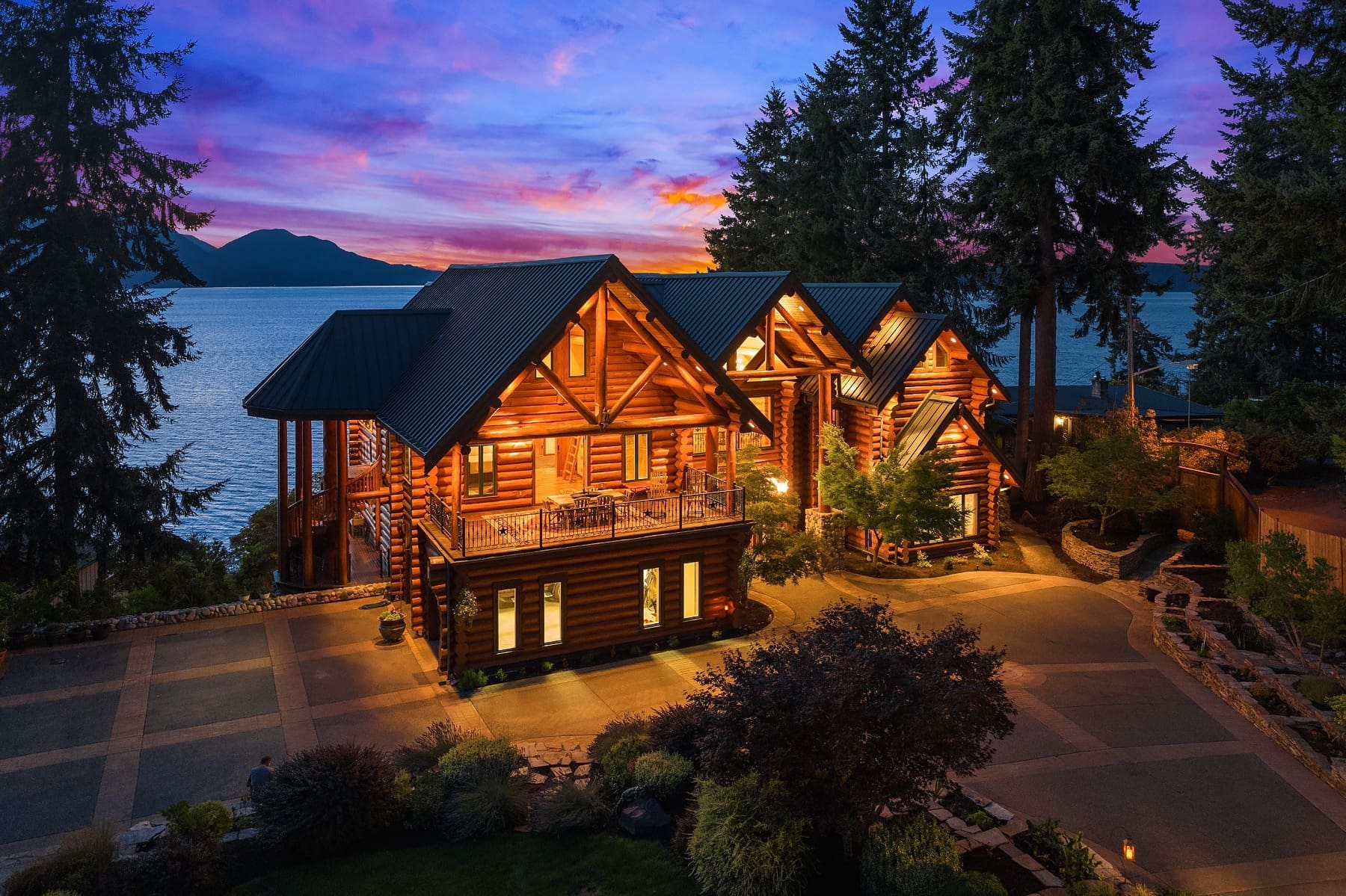 A large, illuminated log cabin with multiple gabled roofs stands near a body of water at dusk. The sky is painted with vibrant hues of purple and pink. Tall evergreen trees frame the house, and the well-lit driveway leads to the serene waterfront.