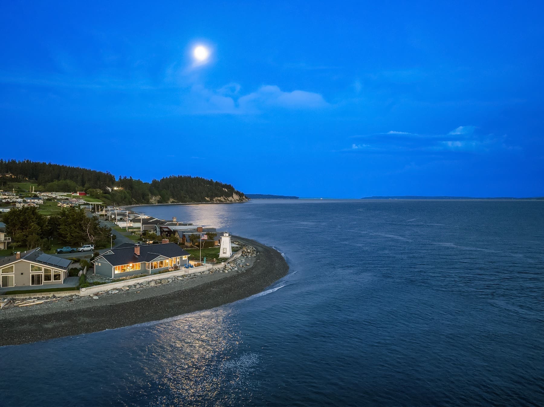 A serene coastal scene at dusk with the moon shining brightly in a clear blue sky. A lighthouse and houses are situated at the edge of a gently curving shoreline. The calm waters of the sea stretch out to the horizon with lush greenery in the background.