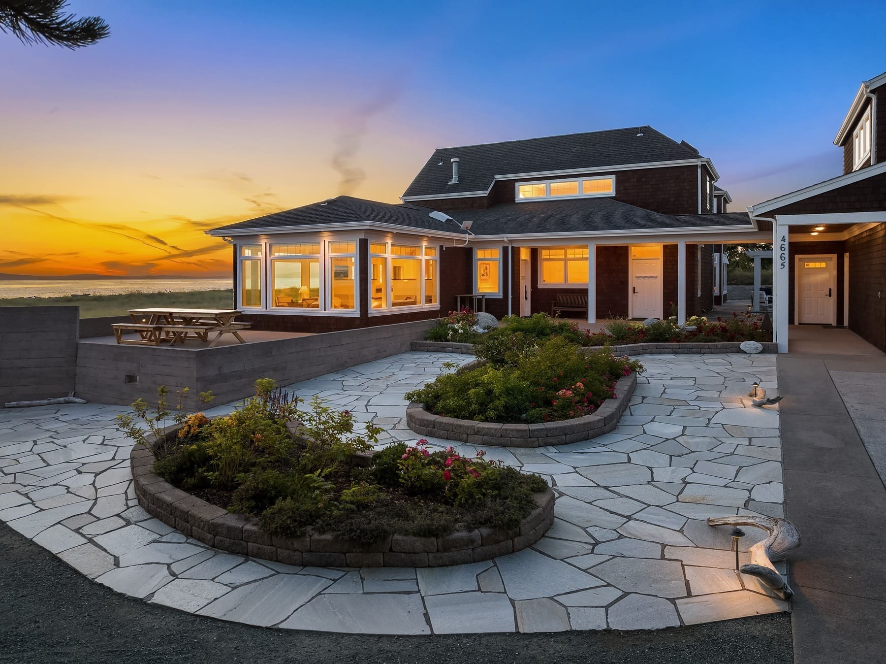 A modern house at sunset with a spacious outdoor patio. The patio features a stone-paved area with raised flower beds, outdoor lighting, and a wooden picnic table. Large windows on the house allow views of the interior, and the sky is painted with warm hues.