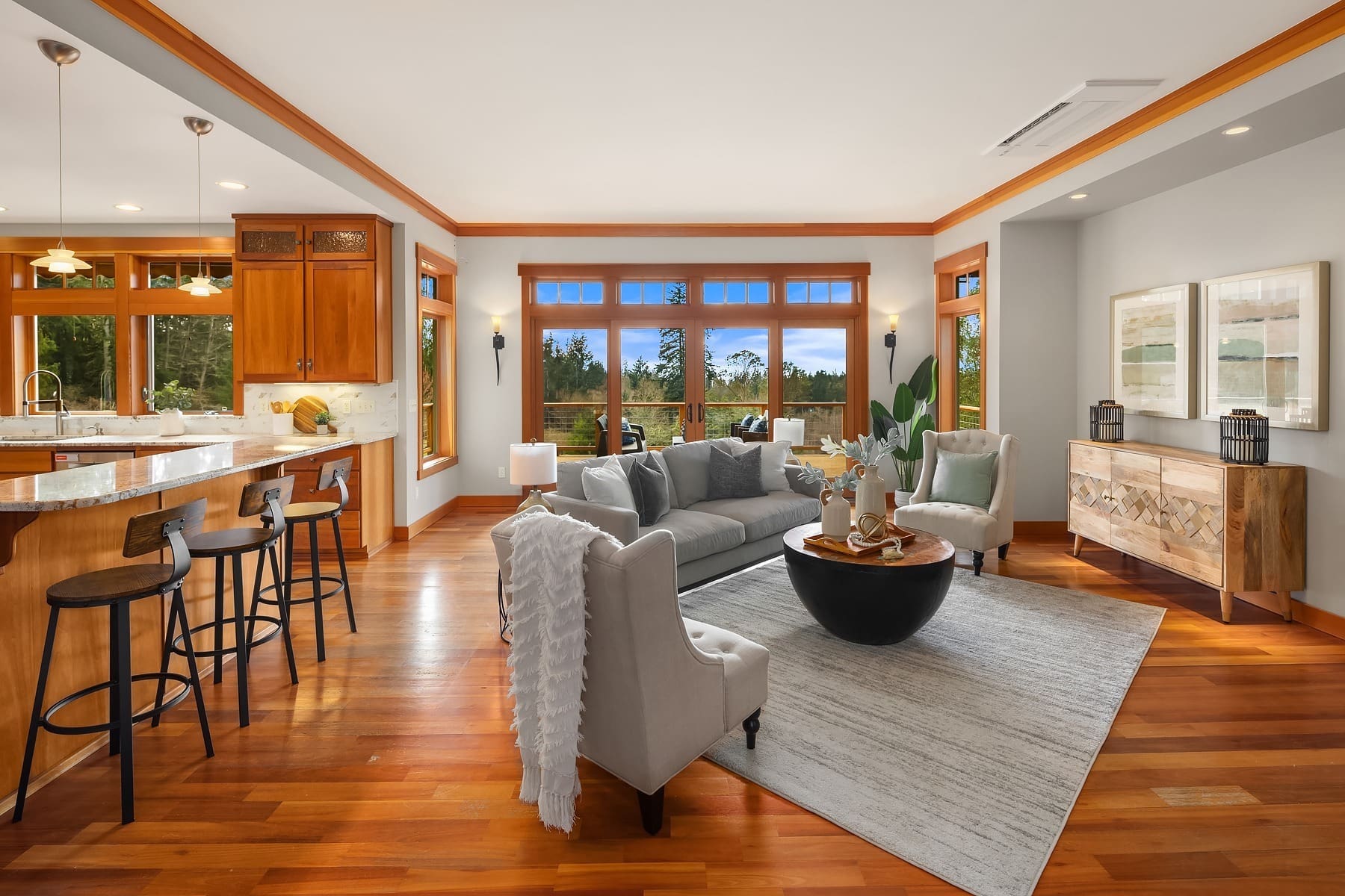 A modern, open-concept living room with hardwood floors, neutral walls, and large windows offering a view of the outdoors. The space includes a gray sofa, armchairs, a coffee table, a rug, a kitchenette with bar stools, and wall-mounted decor and lighting.