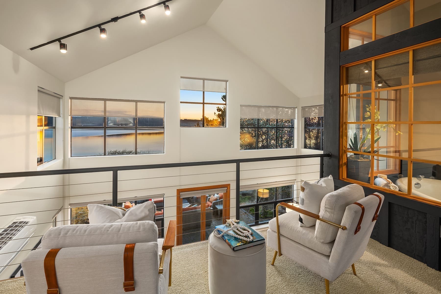 A modern loft space with large windows overlooking a serene body of water at sunset. The area is furnished with two cushioned chairs, a small ottoman, and a cozy throw blanket. Recessed lighting and a metal railing add to the contemporary ambiance.