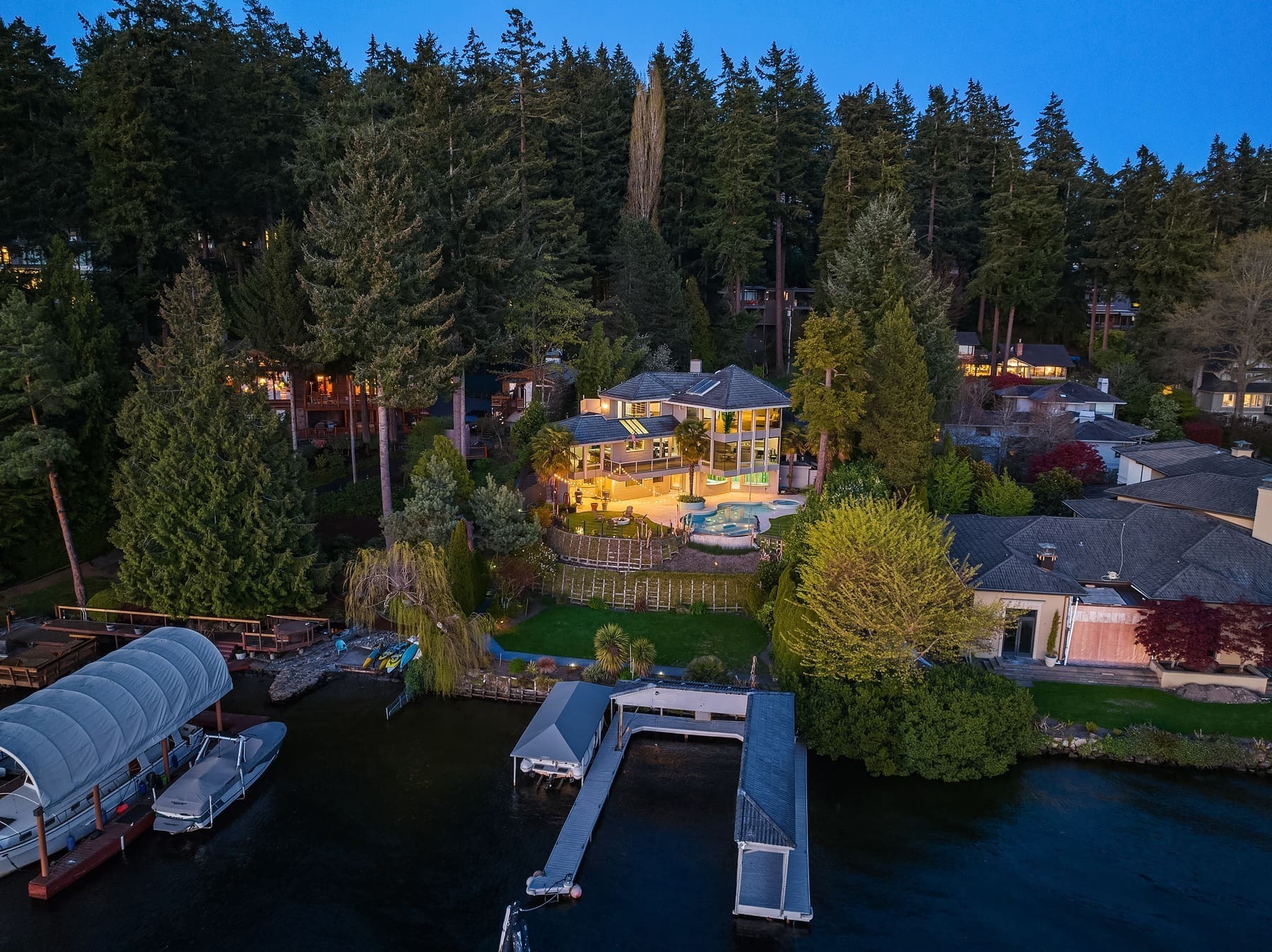 A large, modern house with extensive glass windows sits among tall trees, illuminated in the evening. A swimming pool and landscaped garden are visible. The property is adjacent to a calm lake with docks and boats. Other houses and lush greenery surround the area.