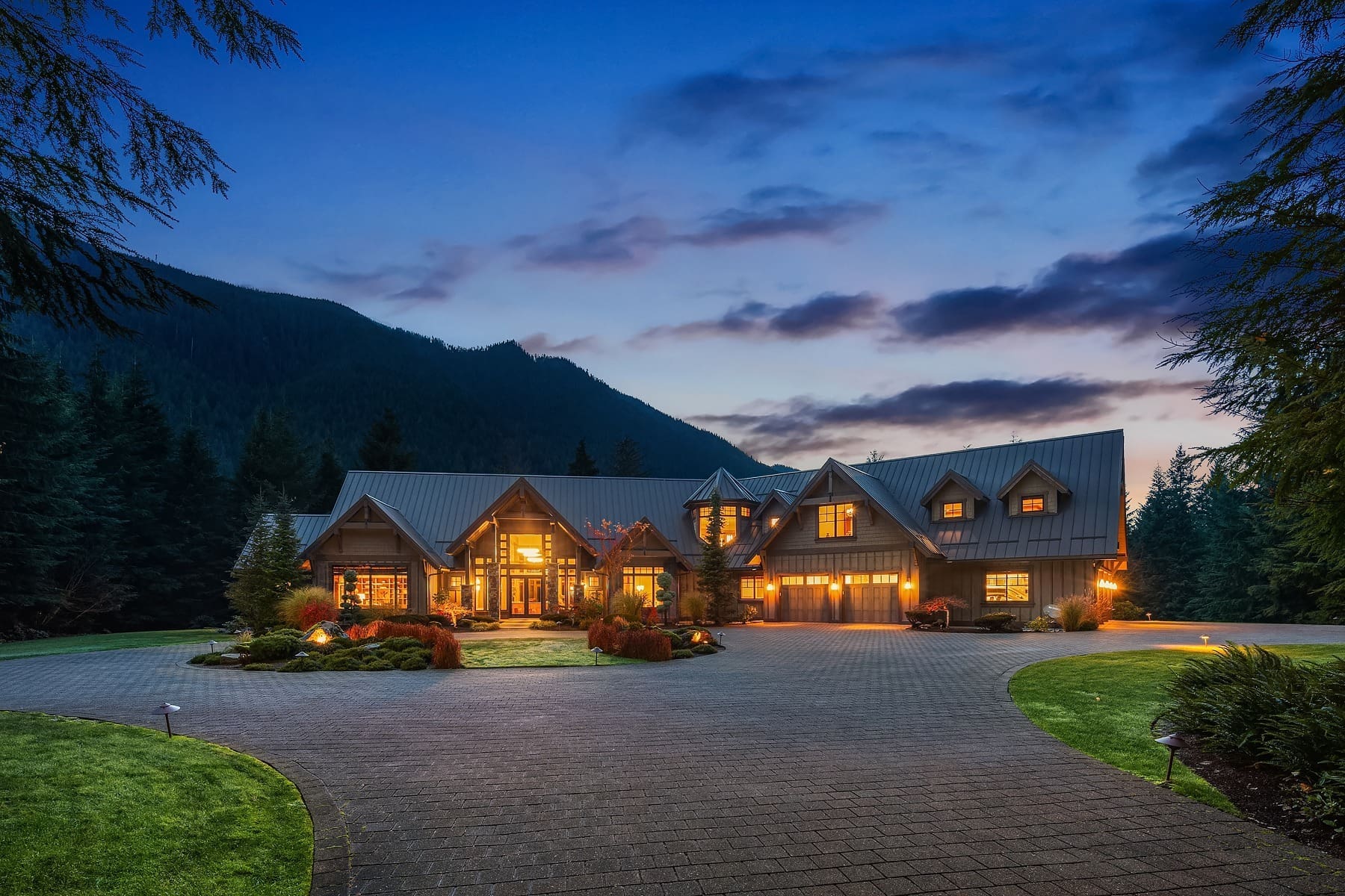 A large, warmly lit wooden house with multiple peaked roofs sits in the twilight amidst lush greenery. A circular driveway fronts the house, with trees and rolling mountains visible in the background under a partly cloudy sky.