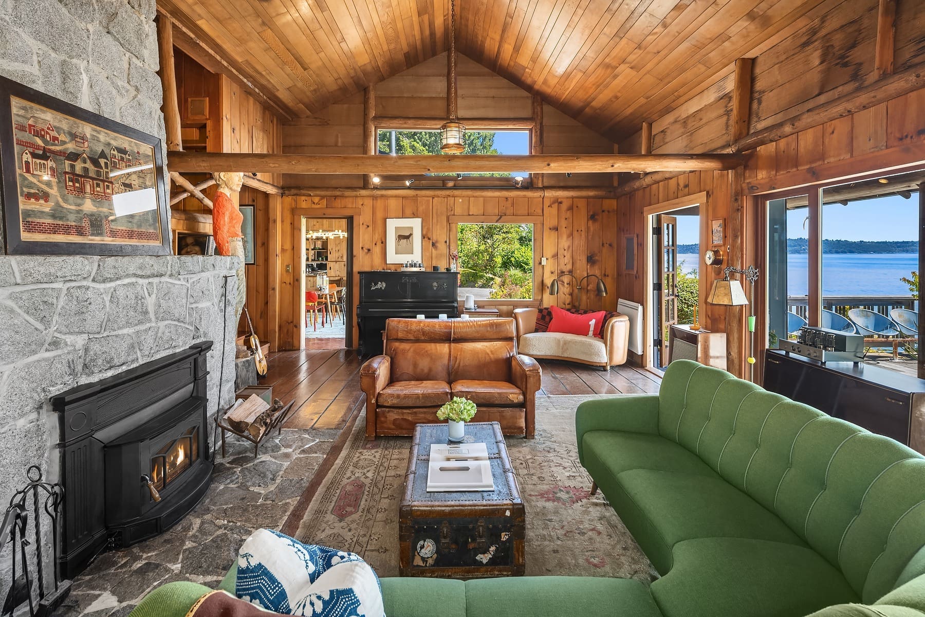 A cozy, rustic living room with wooden walls and ceilings. A green couch, leather chairs, and a stone fireplace create a warm atmosphere. Large windows and glass doors offer a view of a lake. A piano is in the corner, and a patterned rug covers the wooden floor.