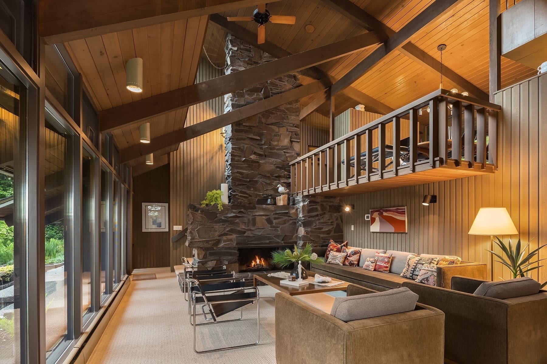 A cozy living room with high wooden ceilings and large windows showcasing a lush green view. The room features a stone fireplace, modern furniture including sofas and chairs, and a small loft area above. Warm lighting and decorative accents add to the inviting atmosphere.