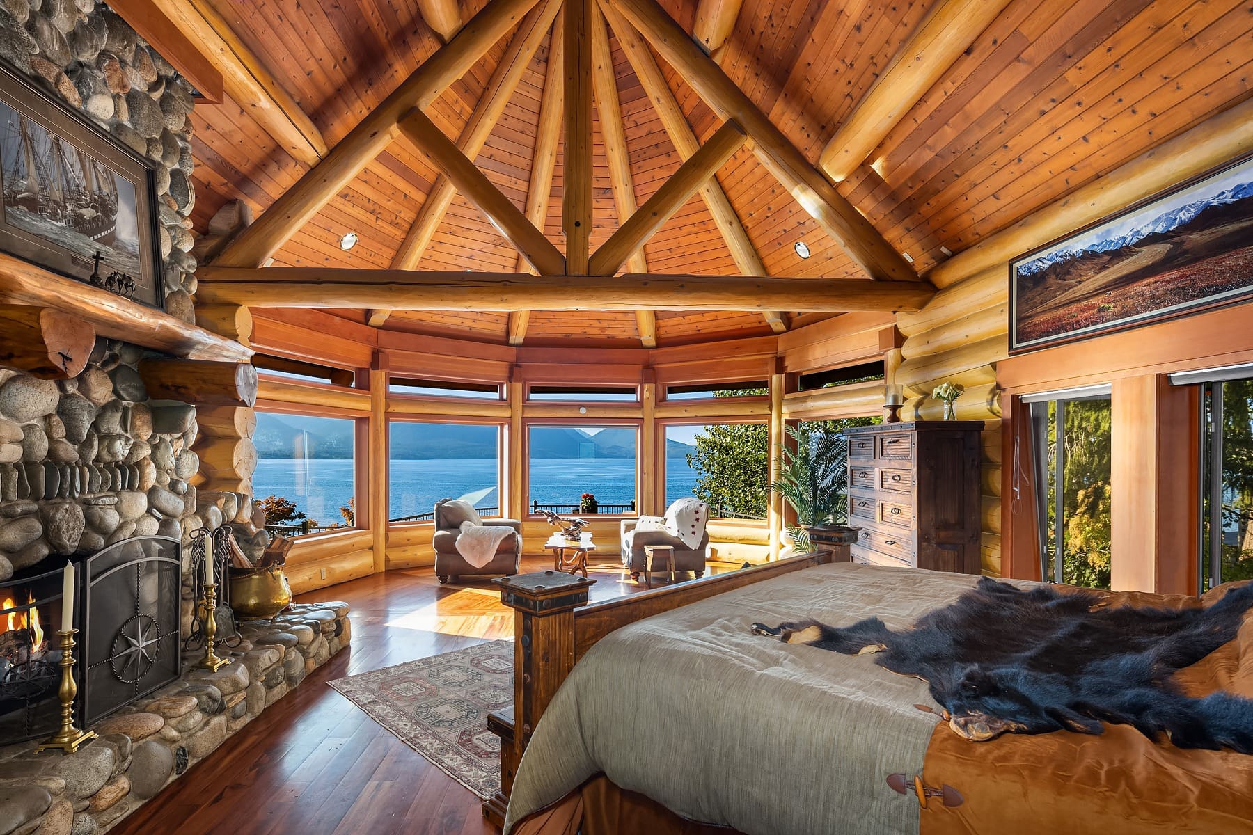 A rustic bedroom with a vaulted wooden ceiling, stone fireplace, and large windows offering a panoramic view of a lake. The room has a large bed with a fur throw, a sitting area with chairs, and various decor items, creating a cozy, cabin-like atmosphere.