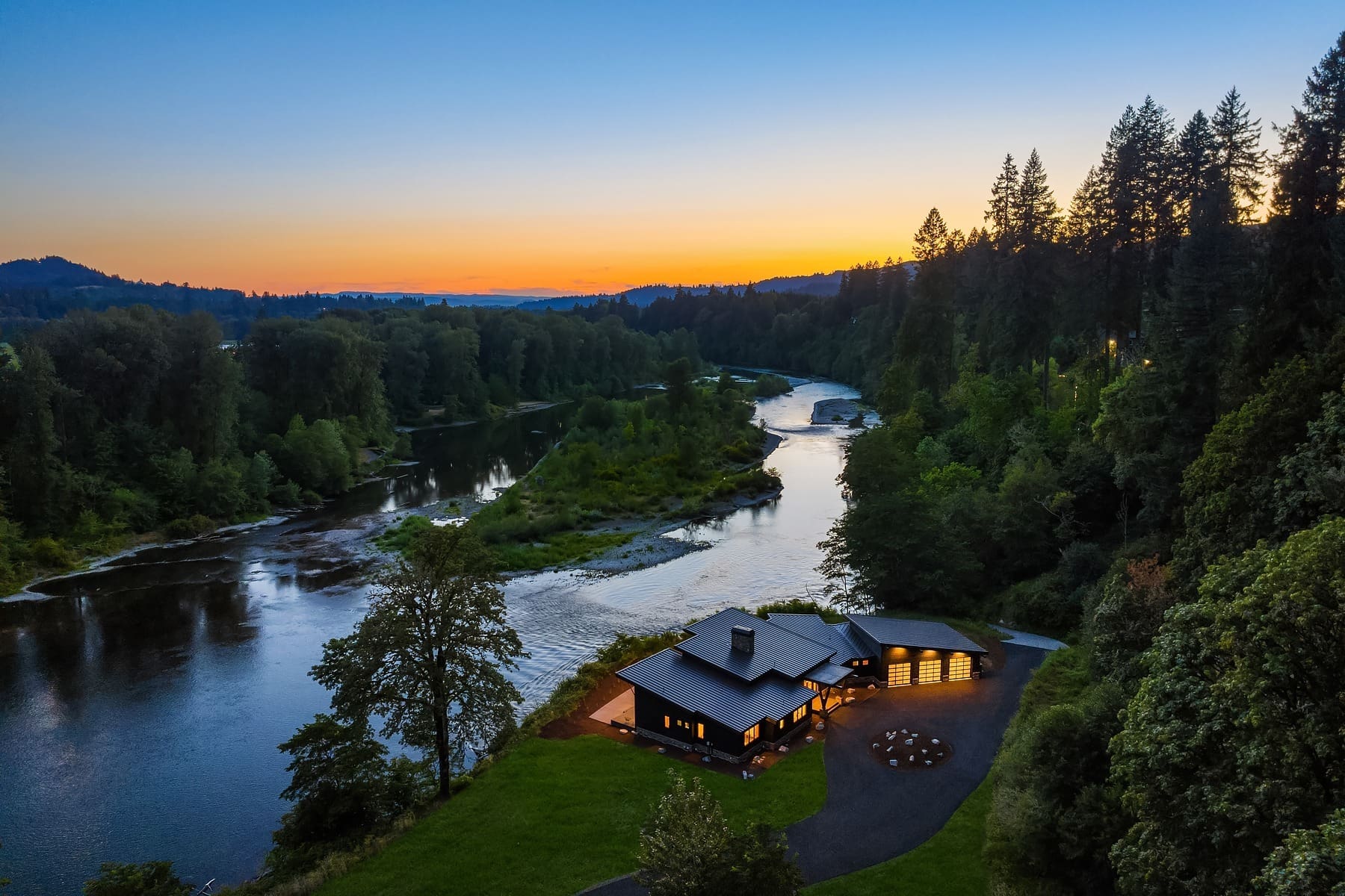 A modern house with large windows sits beside a river in a lush, forested area at sunset. The sky is a gradient of orange to blue and the surrounding trees and landscape are reflected in the calm river. The house is lit warmly, adding to the tranquil atmosphere.