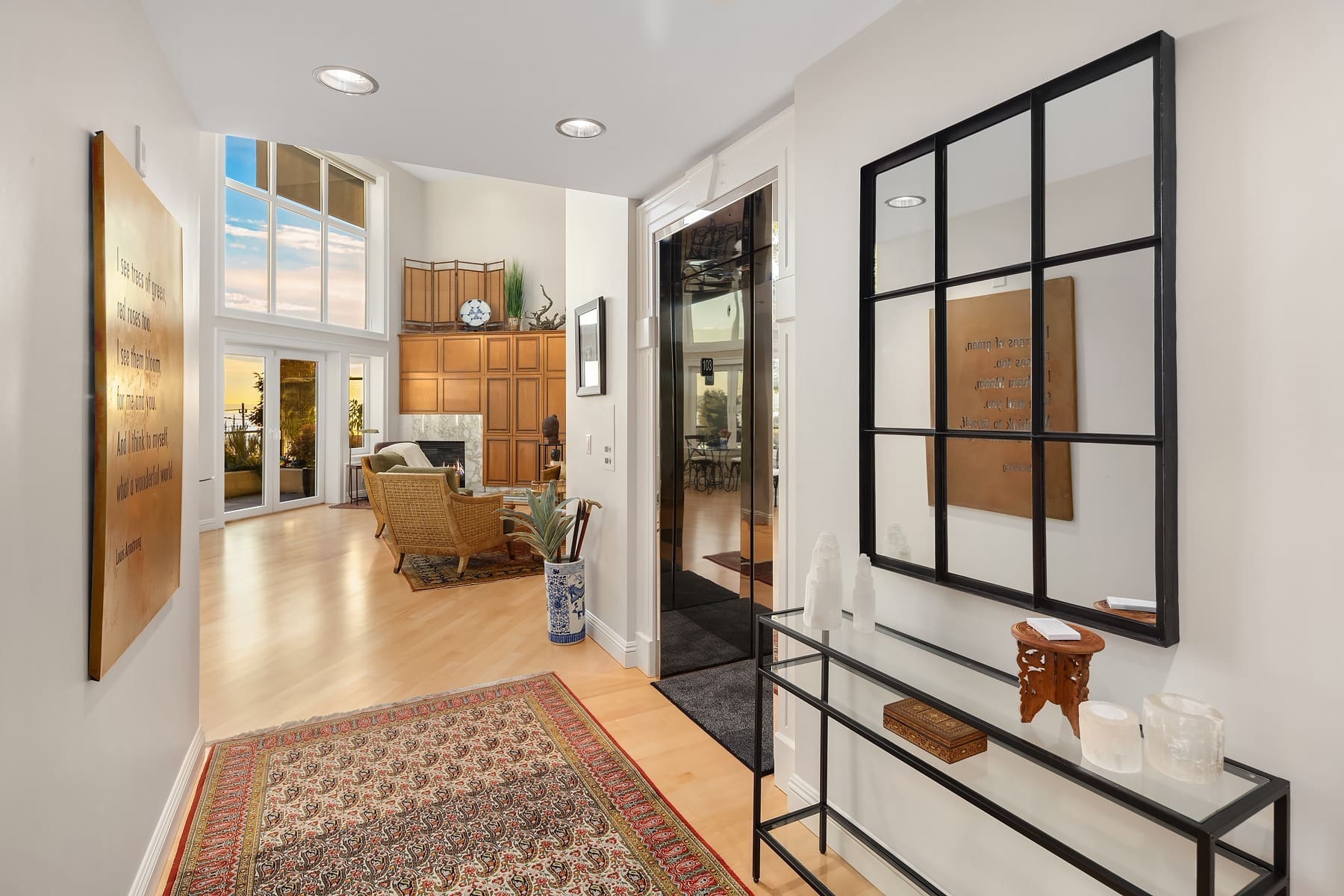 A modern home interior with a bright, open hallway. The hallway features a red patterned rug, wall art, a black-framed mirror, and a glass console table with decor. The hallway leads to a spacious living area with large windows, wooden cabinets, and cozy seating.