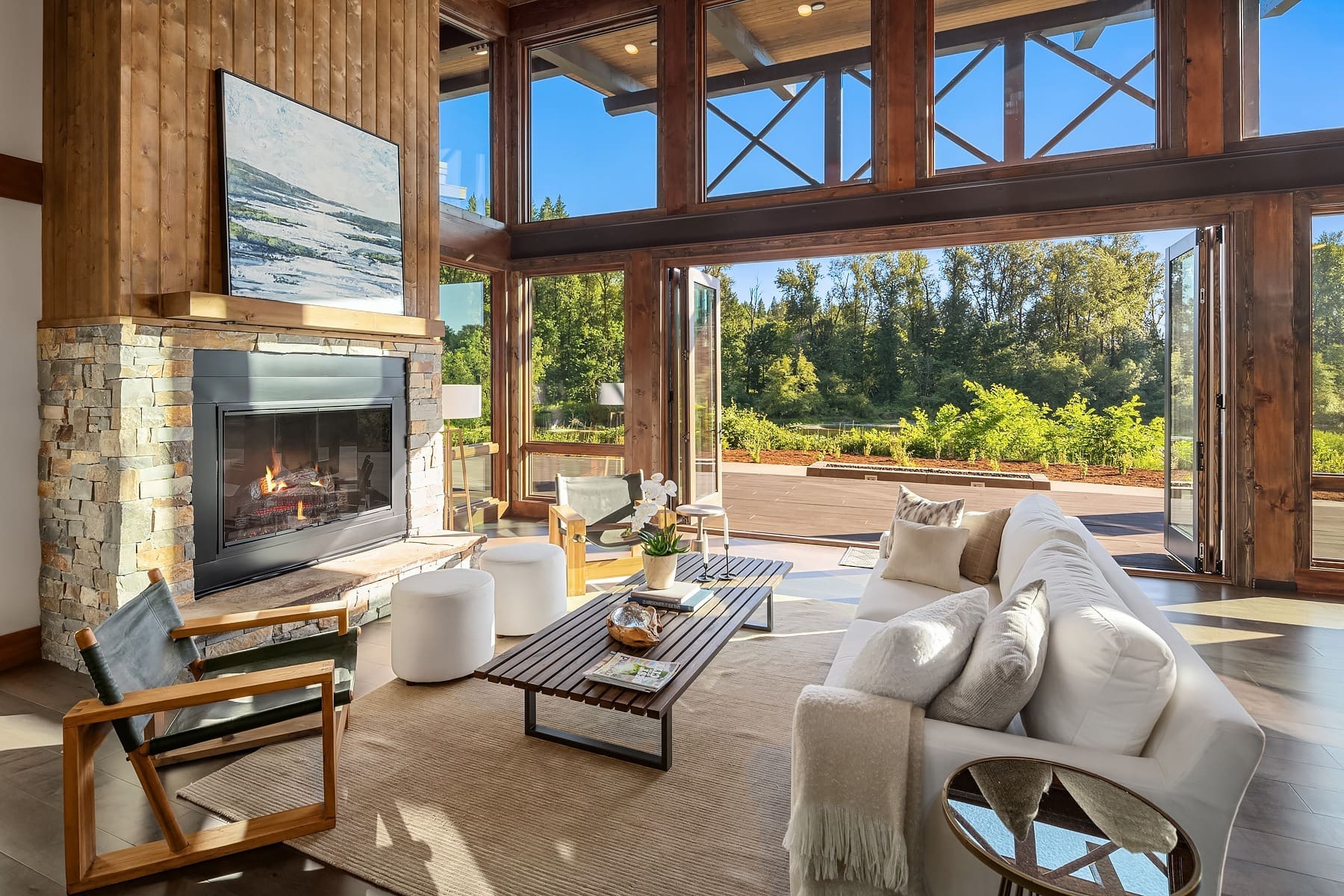 A spacious living room with large windows and abundant natural light. Featuring a modern fireplace on a stone wall, cozy furniture including a white sofa, two white ottomans, and a wooden coffee table. Outside, a deck overlooks a lush green landscape.