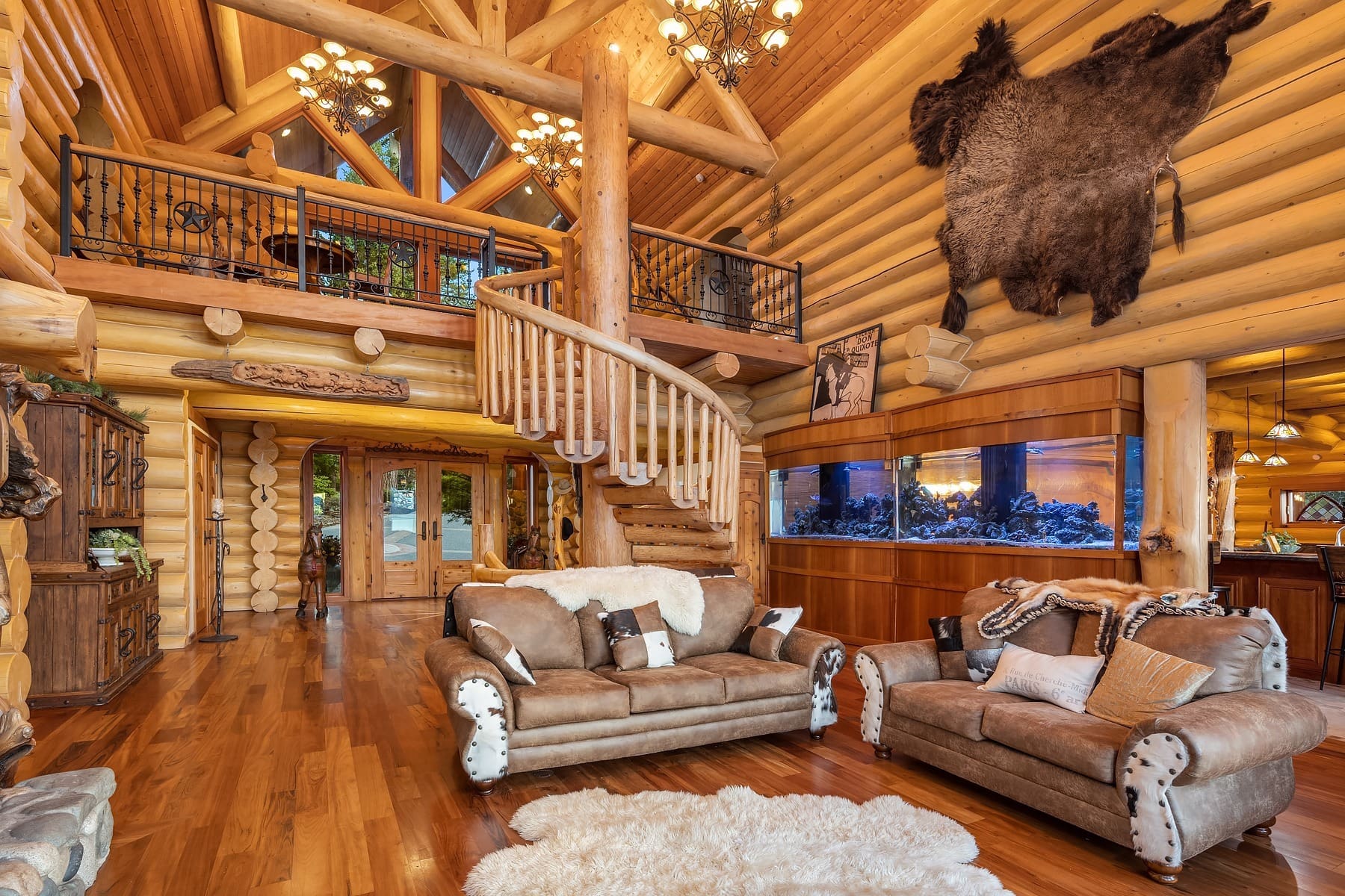 A spacious, rustic log cabin living room featuring plush leather sofas with fur accents, a large spiral staircase, and a mounted animal hide on the wall. An extensive aquarium and wooden balcony railings add to the cozy and natural ambiance.